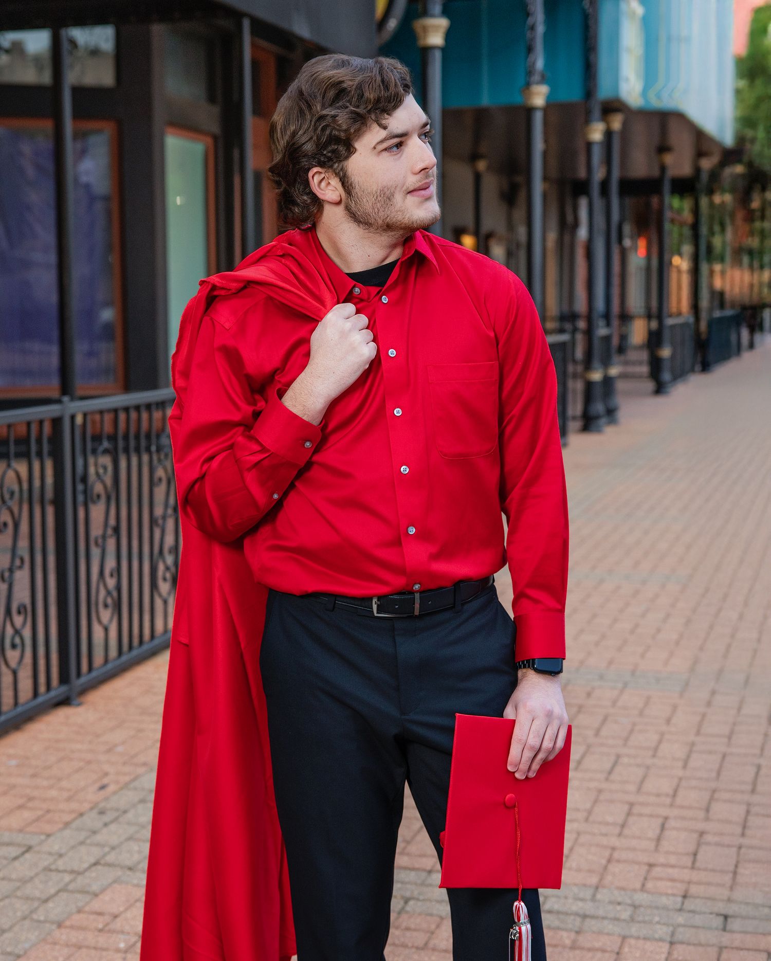 Person in a vibrant red shirt and black pants poses casually against an outdoor storefront walkway.
