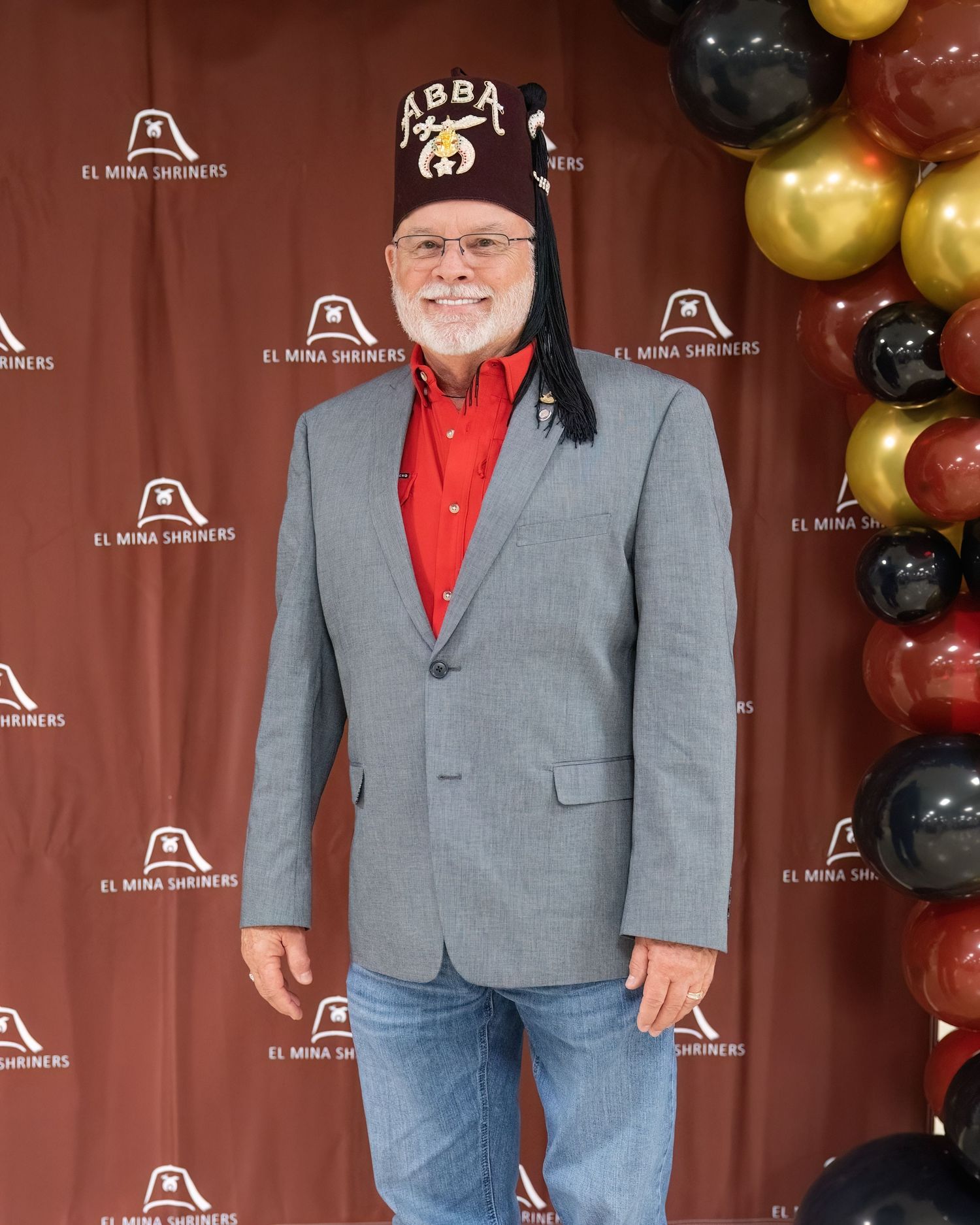 Shriners Gala event with person in a gray suit jacket and red shirt stands against a brown backdrop with gold and black balloons.