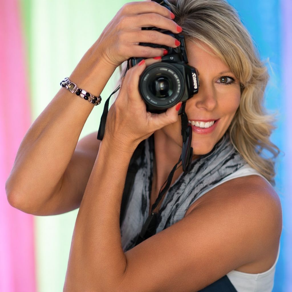 Person holding a camera and smiling against a colorful striped background. Lisa Anderson with LK Anderson Photography
