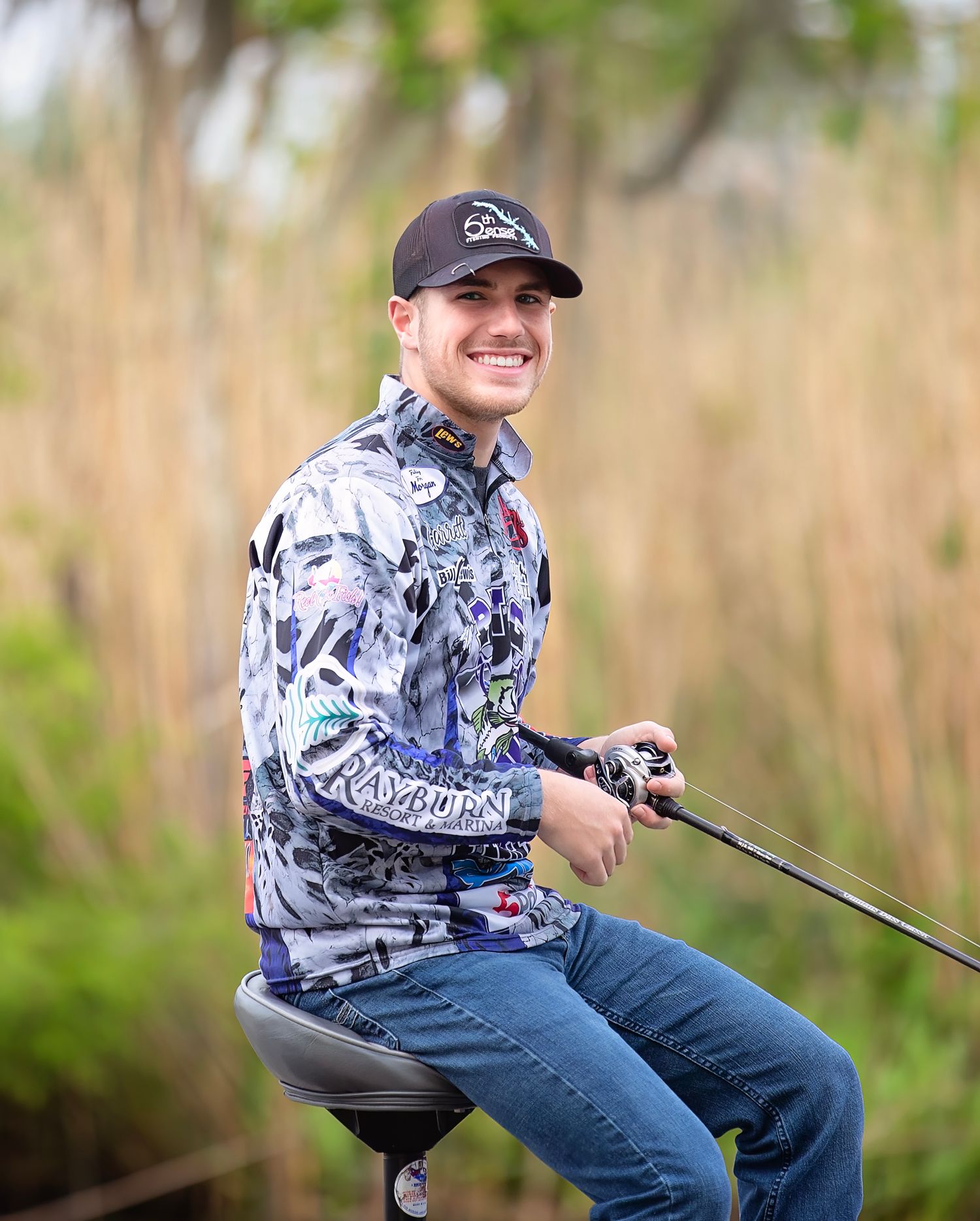 Young man smiling outdoors holding a fishing rod, wearing a patterned shirt and dark cap near tall grass.