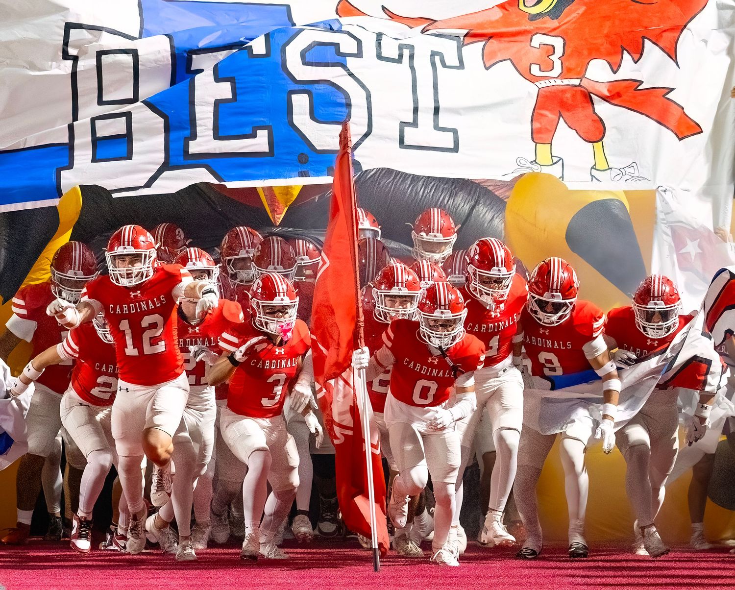 Football team in red jerseys bursts through a banner at a stadium entrance with 'BEST' text visible above.