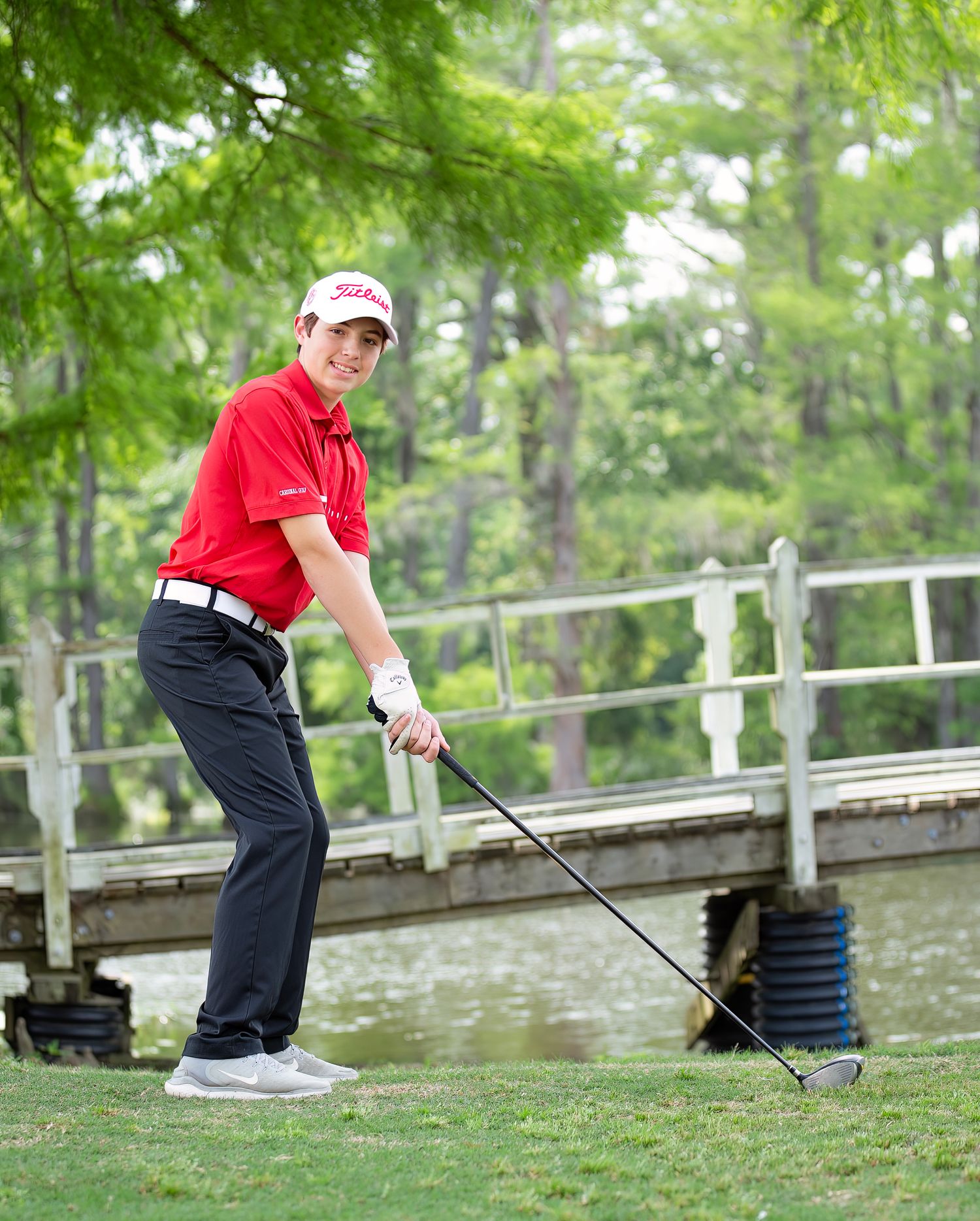 Golfer in red shirt executes a chip shot from the rough near a wooden bridge on a lush golf course.
