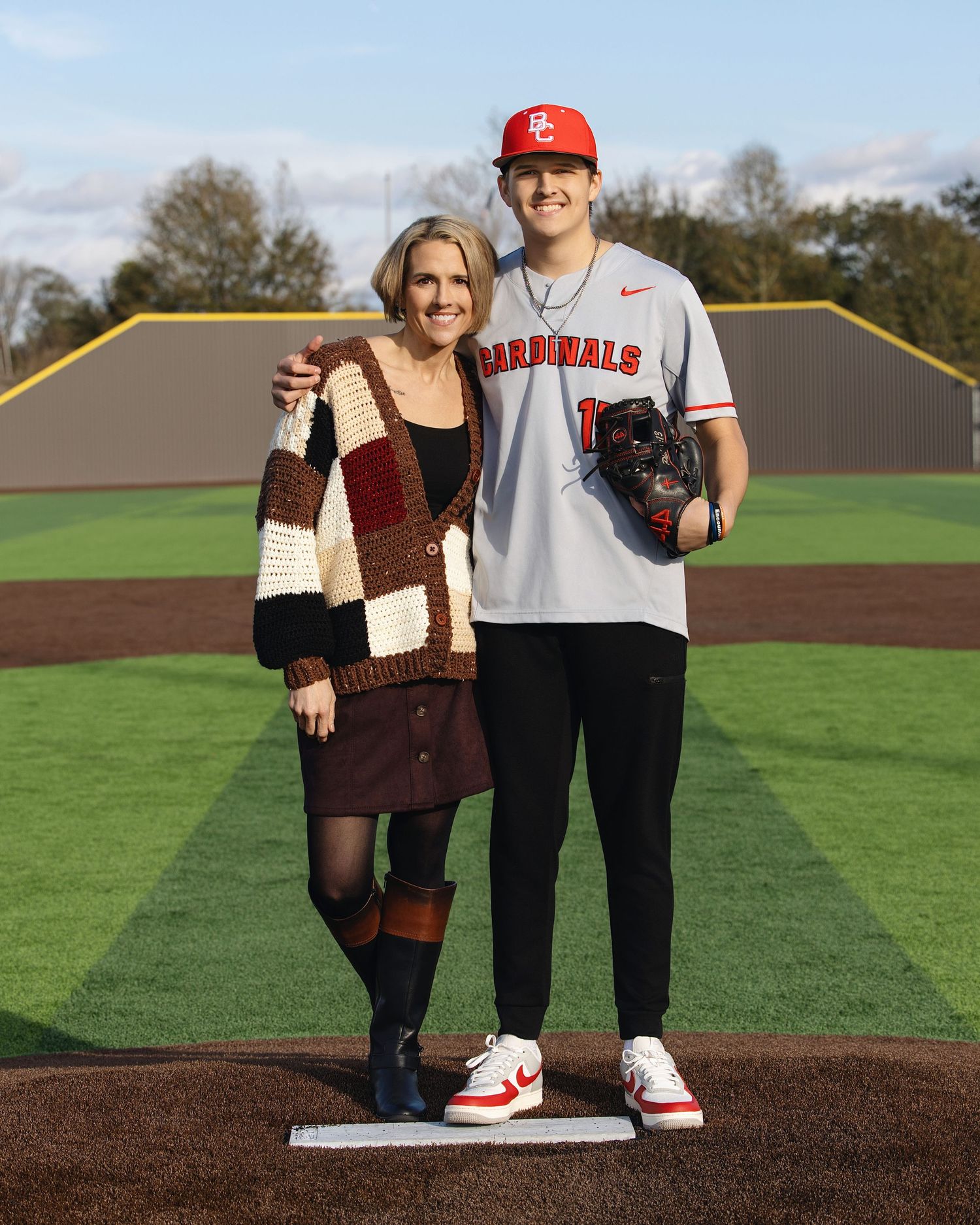 Two people stand together on a baseball field wearing casual clothing and sneakers.