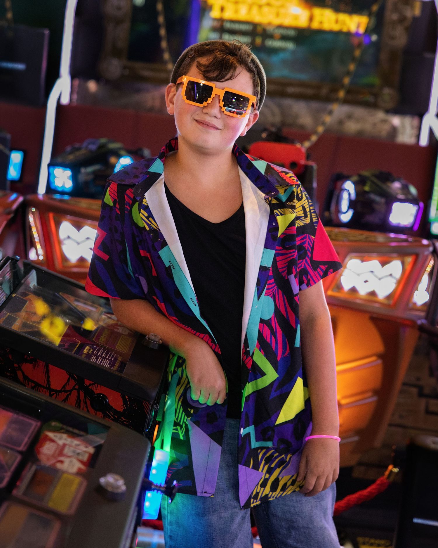 A young boy wearing colorful 80s-style jacket and orange sunglasses poses confidently in a vibrant arcade game room.