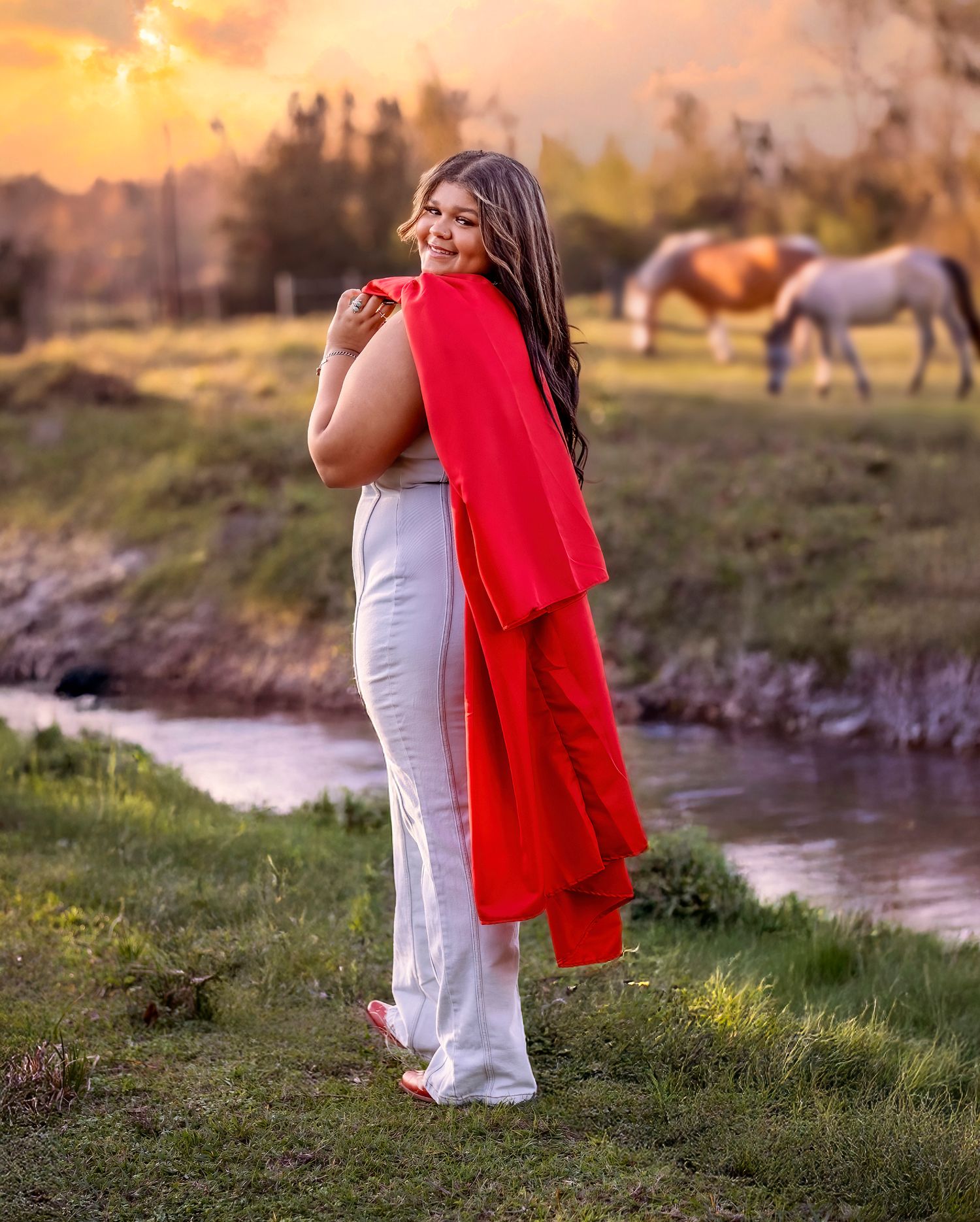 A dreamy sunset portrait in white pants and flowing red shawl beside a stream with horses grazing in the background.