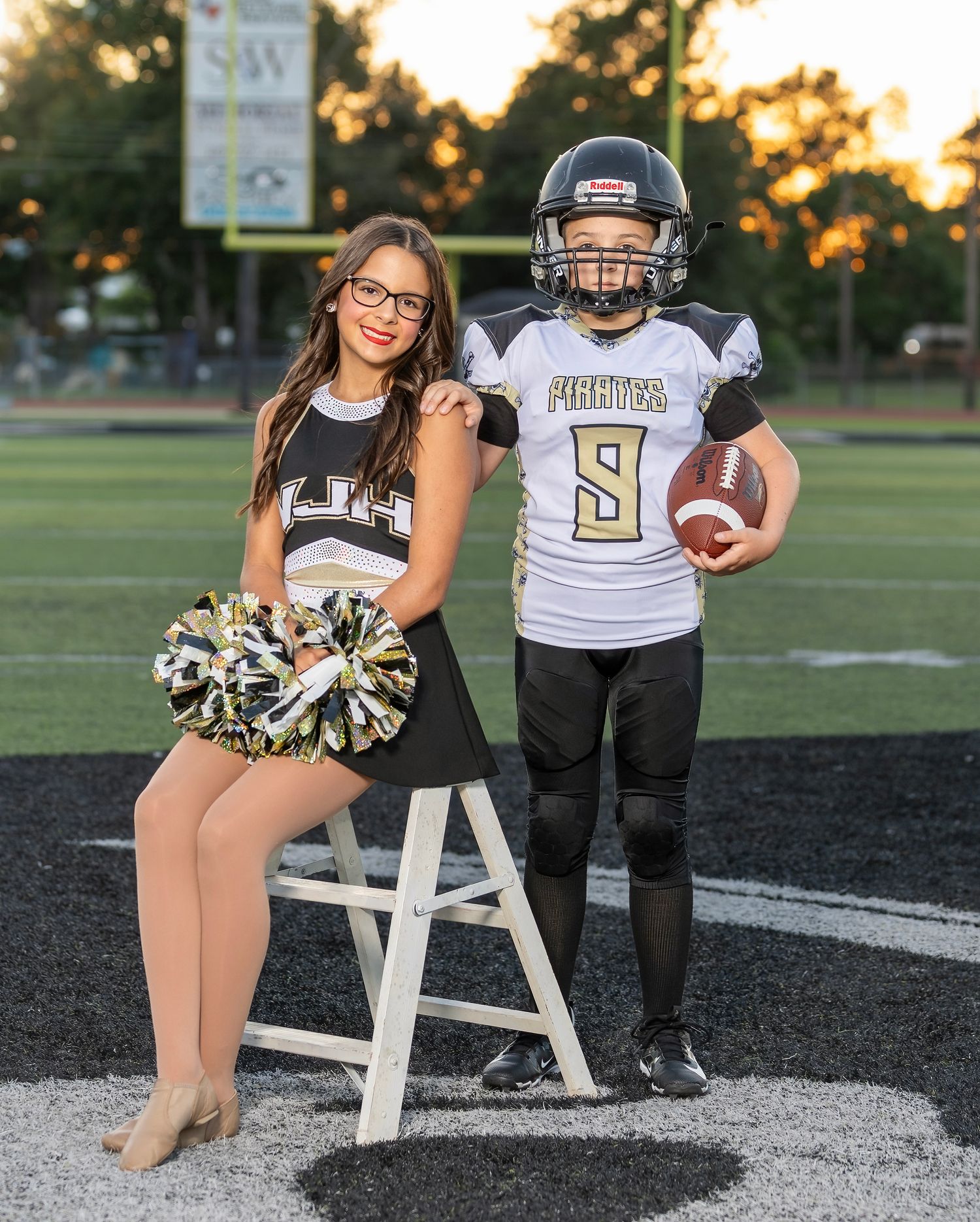 A football player and cheerleader pose together on a football field during sunset for a sports photo session.