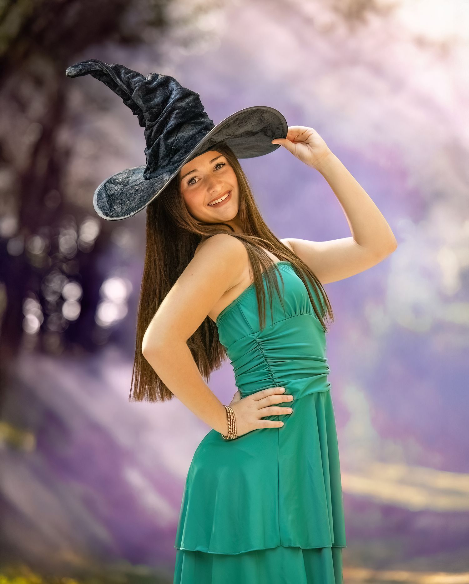 A woman in a green dress and black hat poses playfully in a dreamy outdoor setting with purple tones.