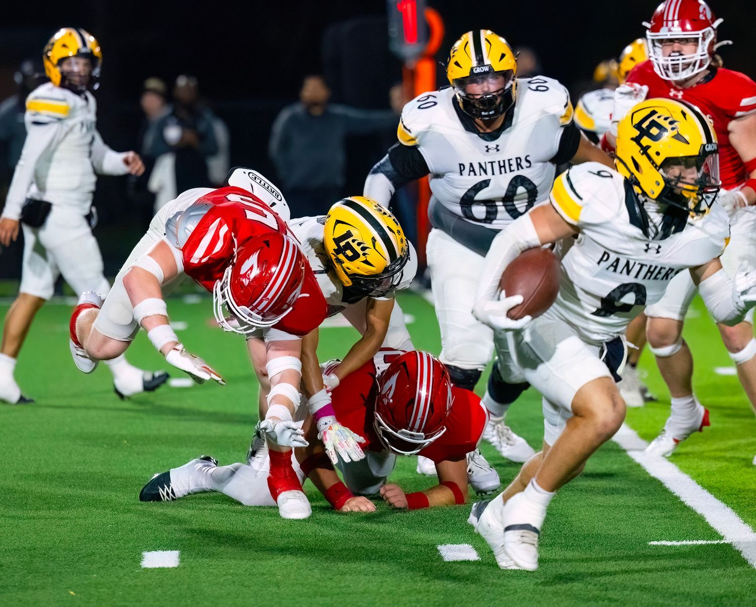 Football players in red and yellow uniforms compete for possession during a ground play on the field.