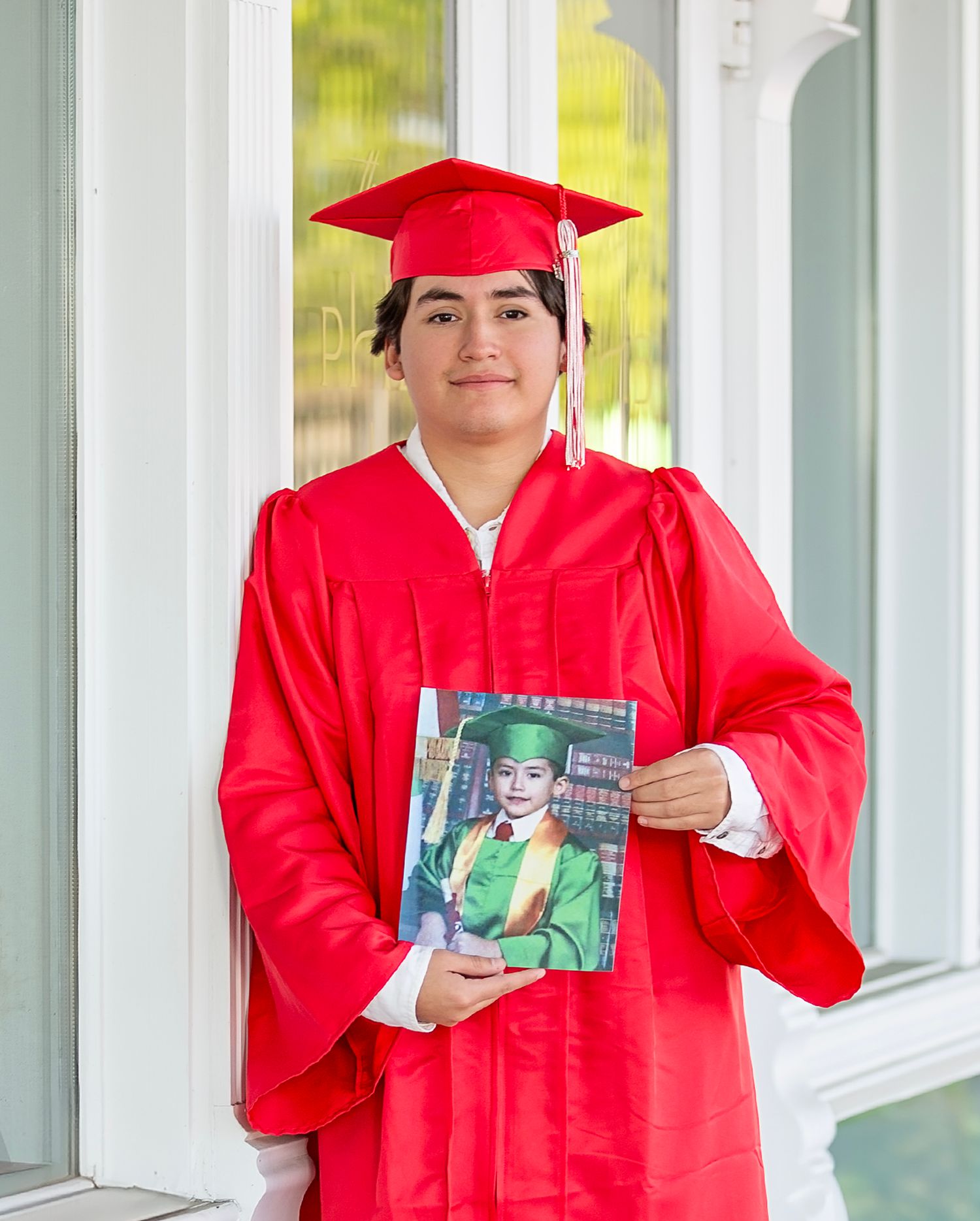 Graduate in bright red cap and gown holds up a framed graduation photo while standing on a white porch.