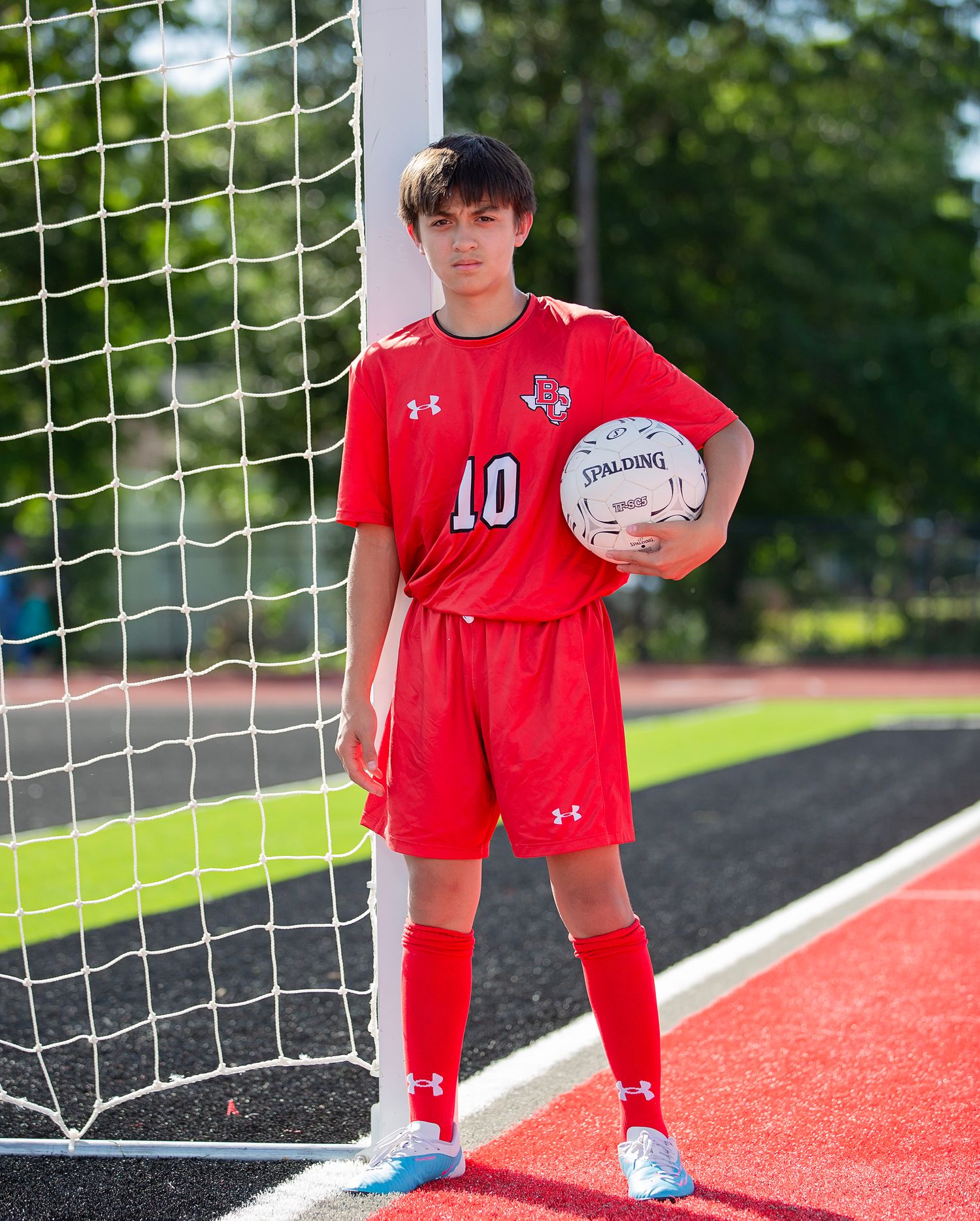 Soccer player in red Under Armour uniform with number 10 poses with ball by goal post on outdoor field.