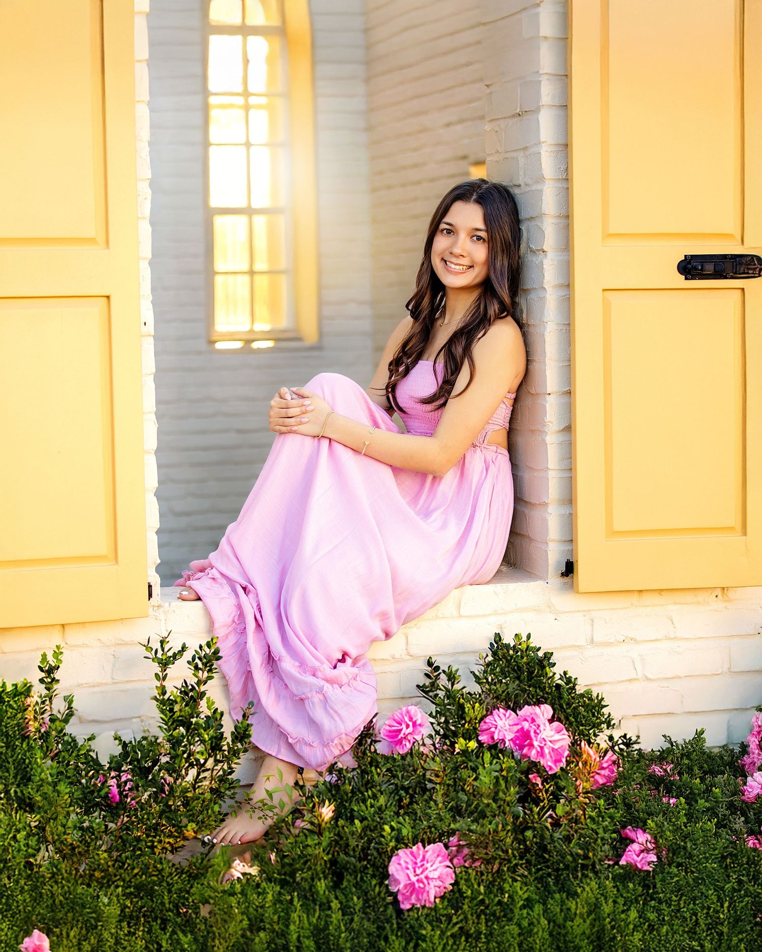 Young woman in pink dress sitting in yellow doorway surrounded by pink flowers and lush greenery.