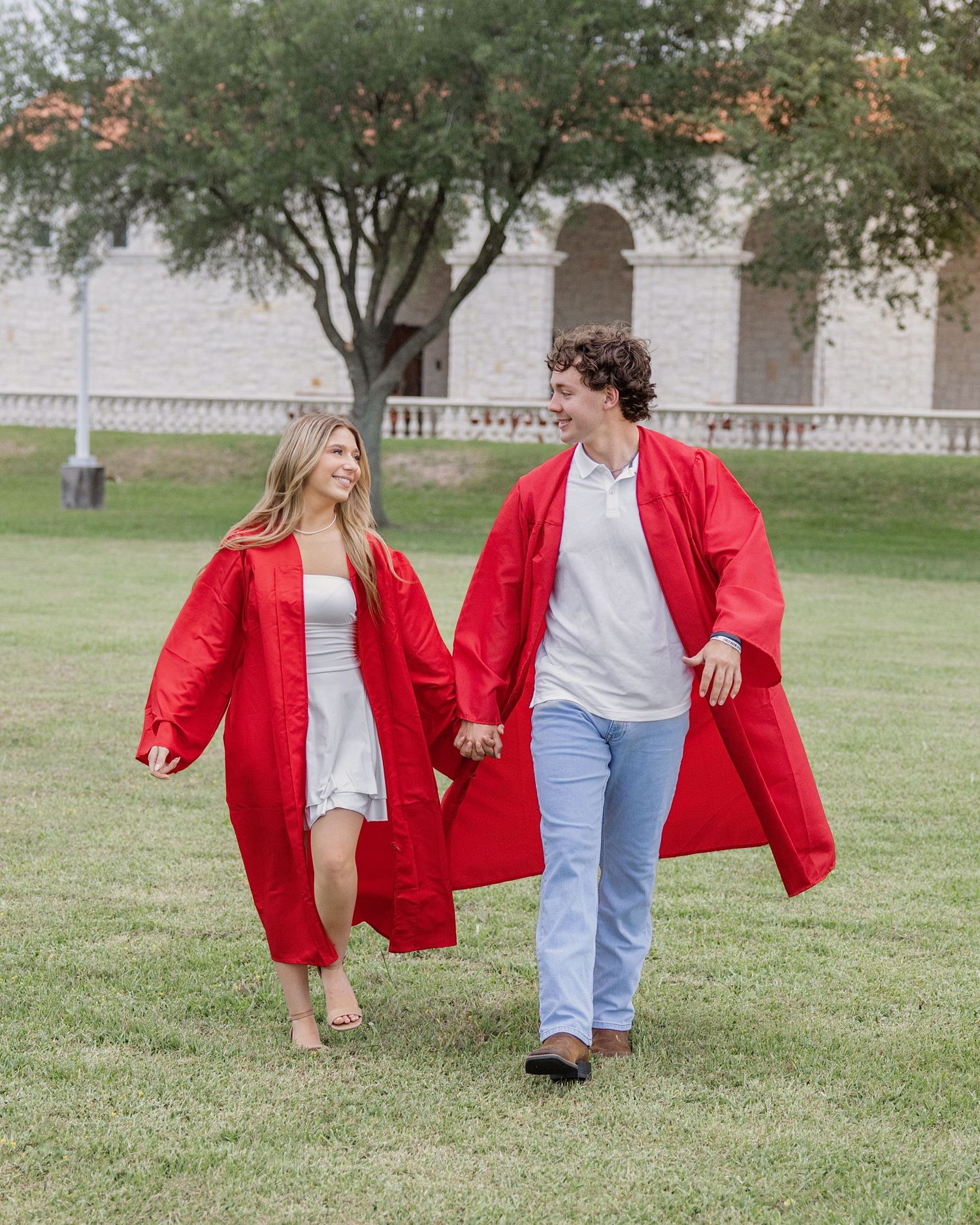 Two graduates in red gowns walk hand in hand on a grassy campus lawn, smiling at each other.