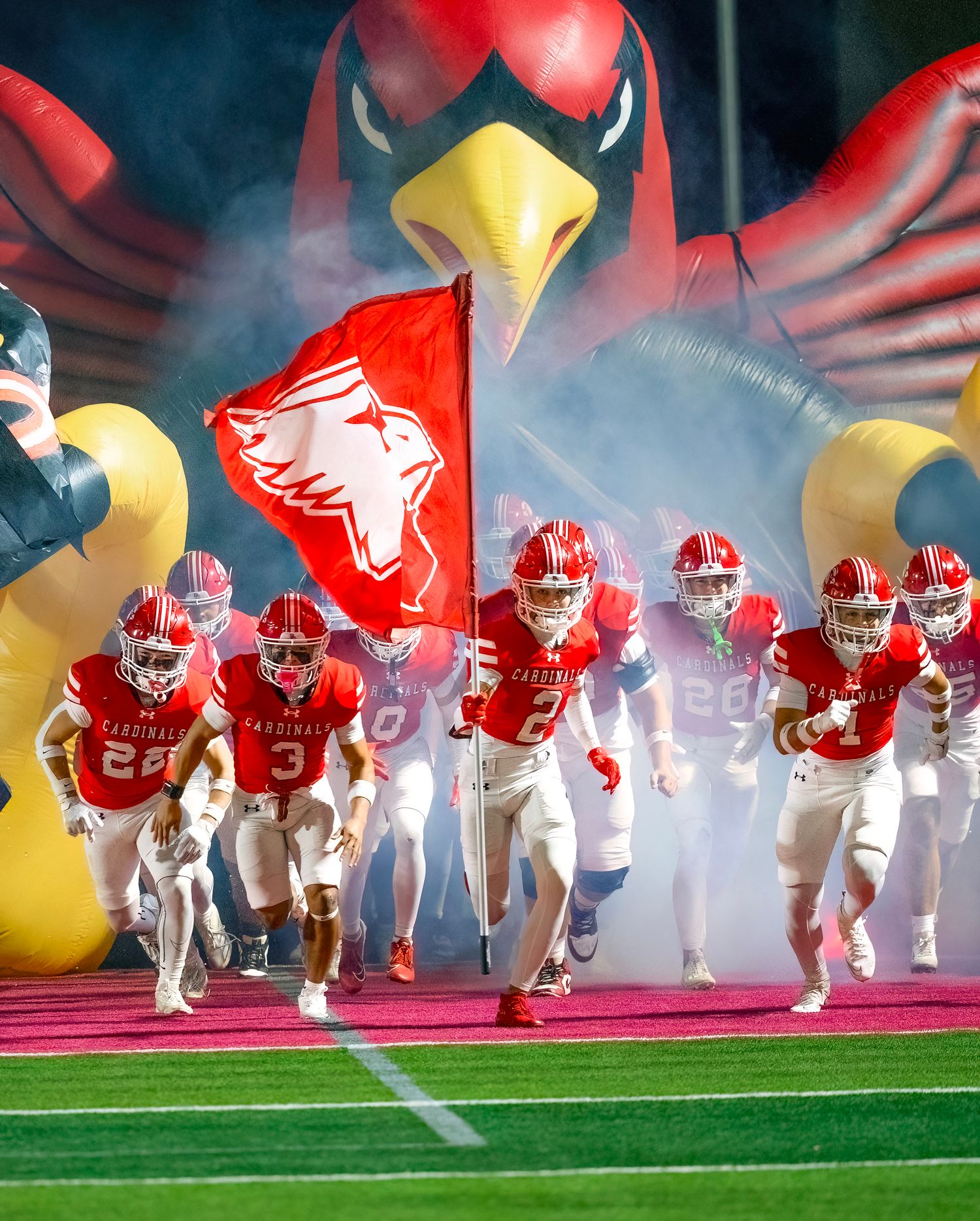 College football players run through smoke onto field led by mascot flag and giant inflatable cardinal mascot head.