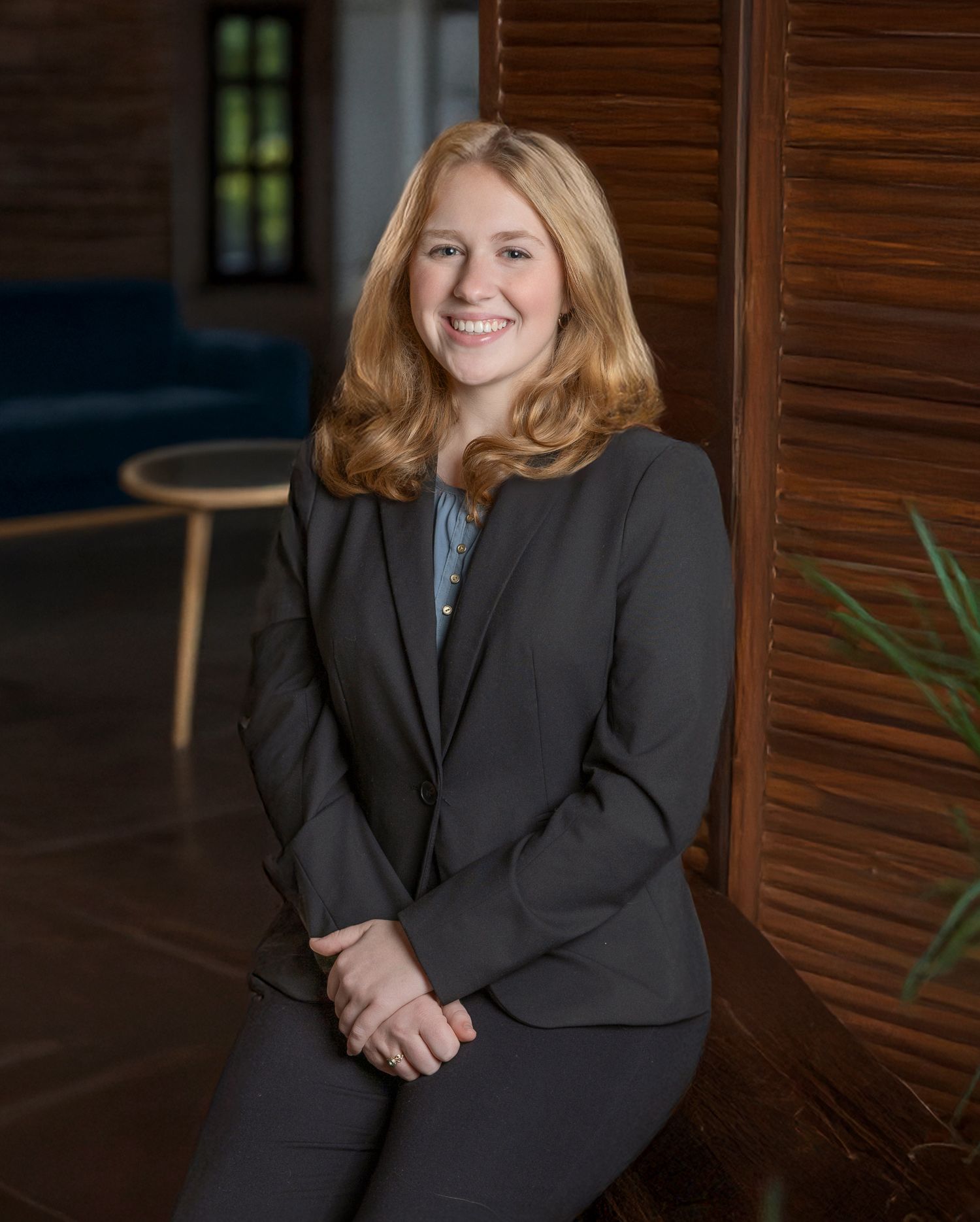 Professional headshot of someone in a dark blazer seated against a wooden wall with warm lighting.