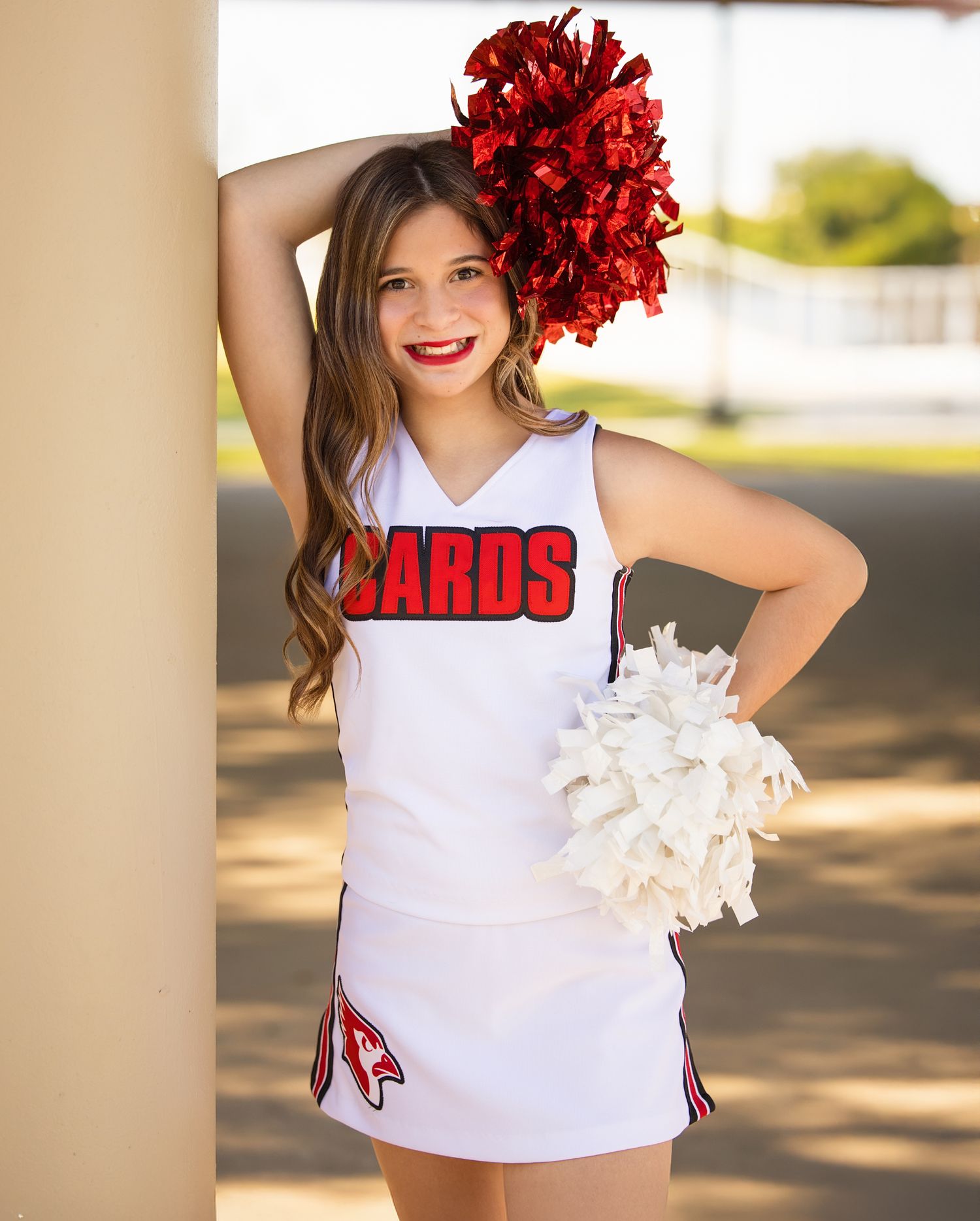 Cheerleader in white uniform top holding red pom poms performs outdoors in bright sunlight.
