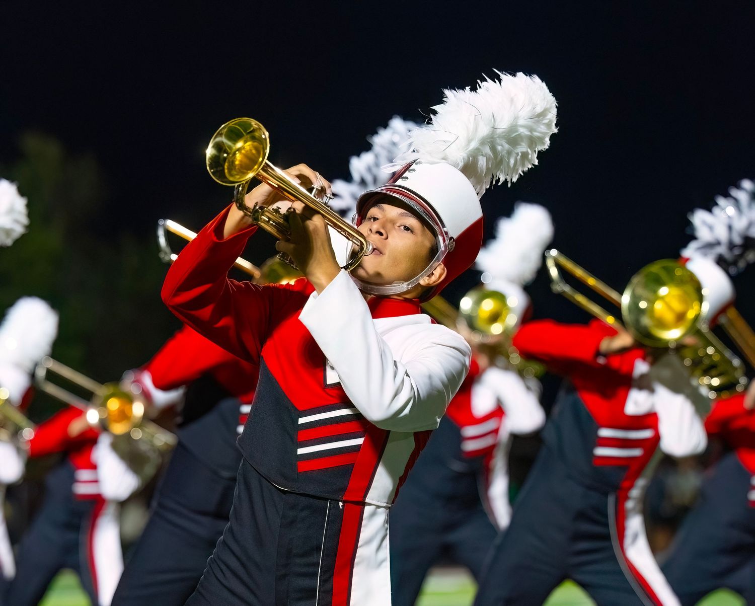 Marching band members in black uniforms perform with brass instruments during a nighttime show.