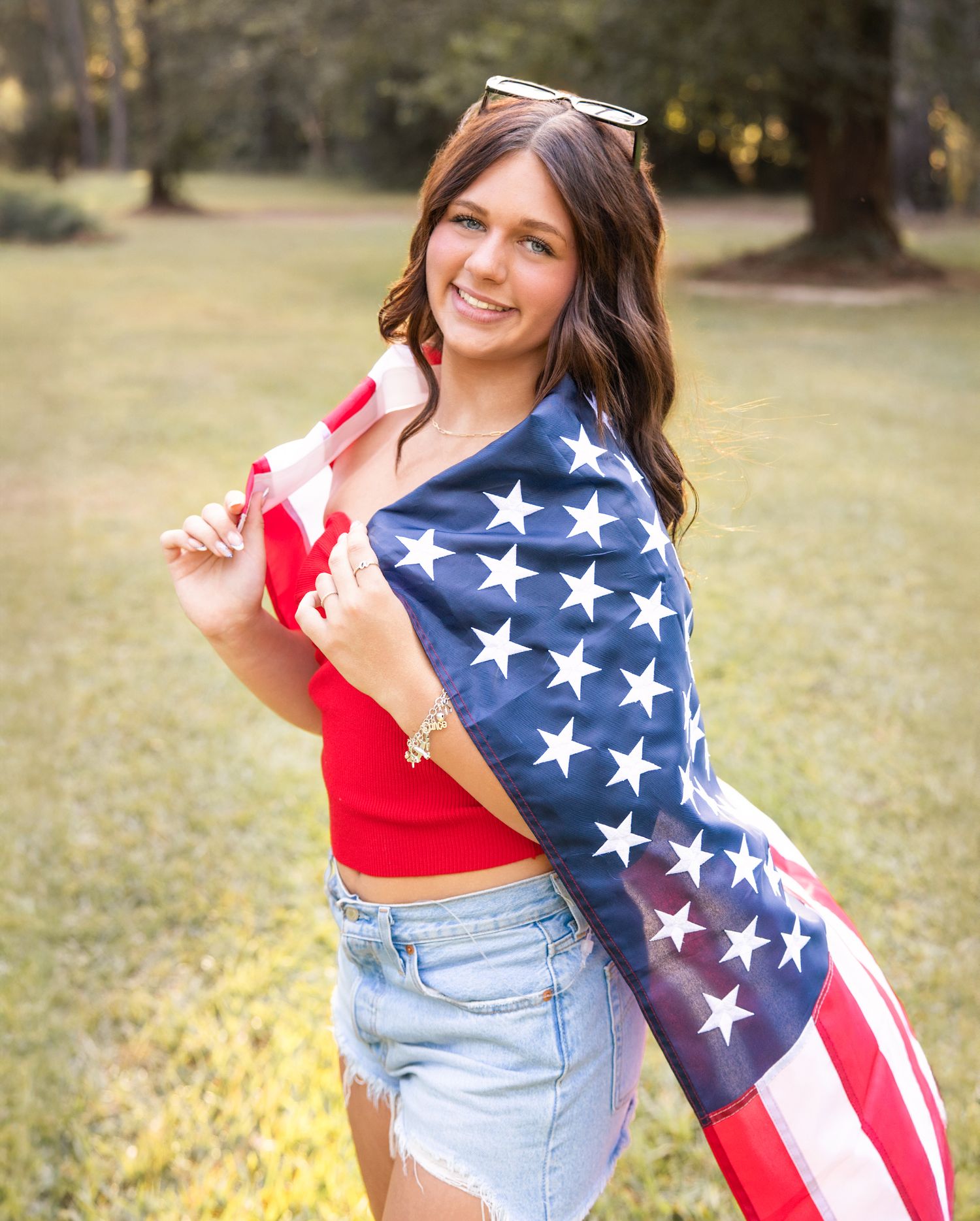 Patriotic outdoor photoshoot featuring American flag and casual summer outfit with red top and denim shorts.