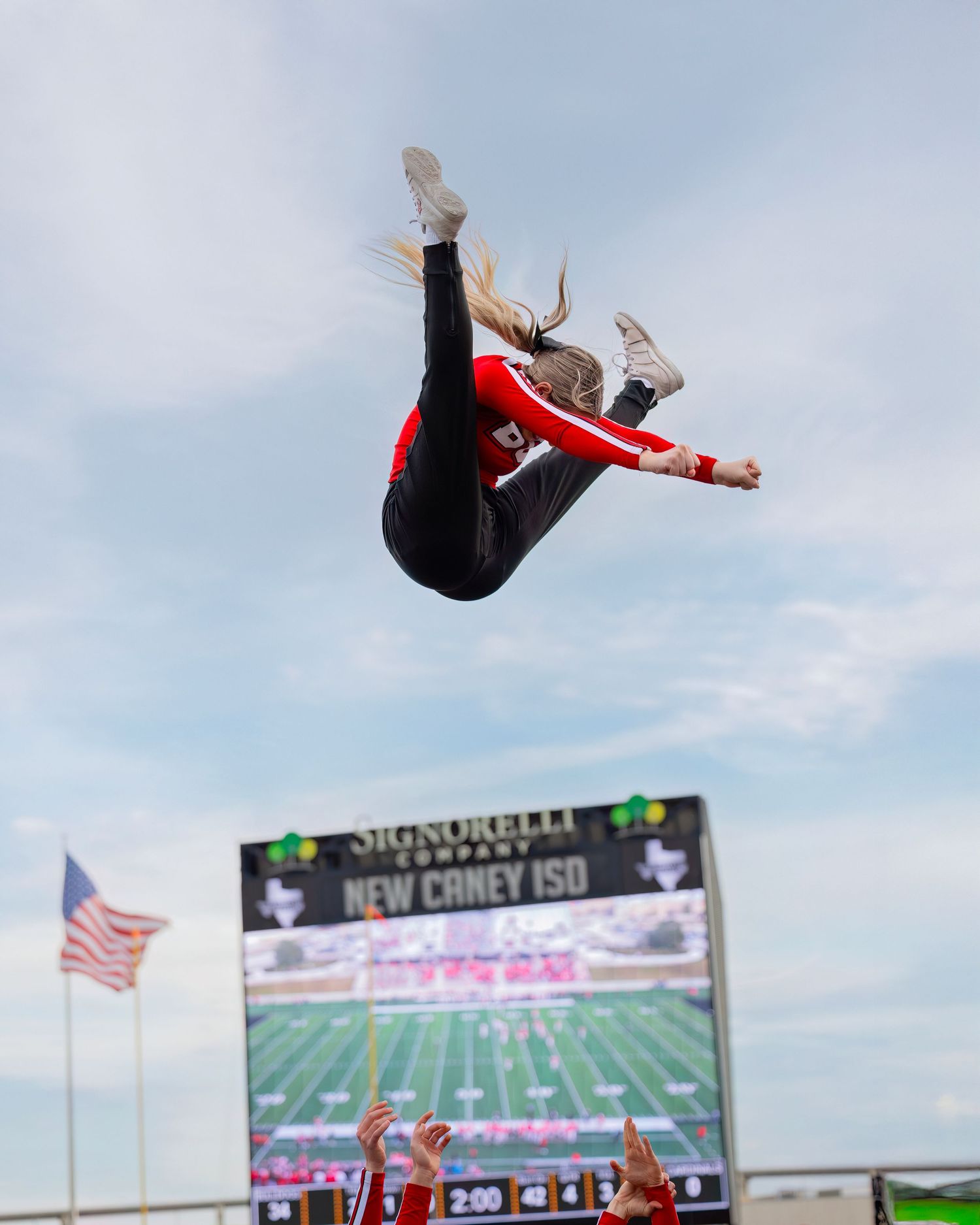 Senior Cheerleader performs aerial backflip against backdrop of stadium scoreboard and blue sky.