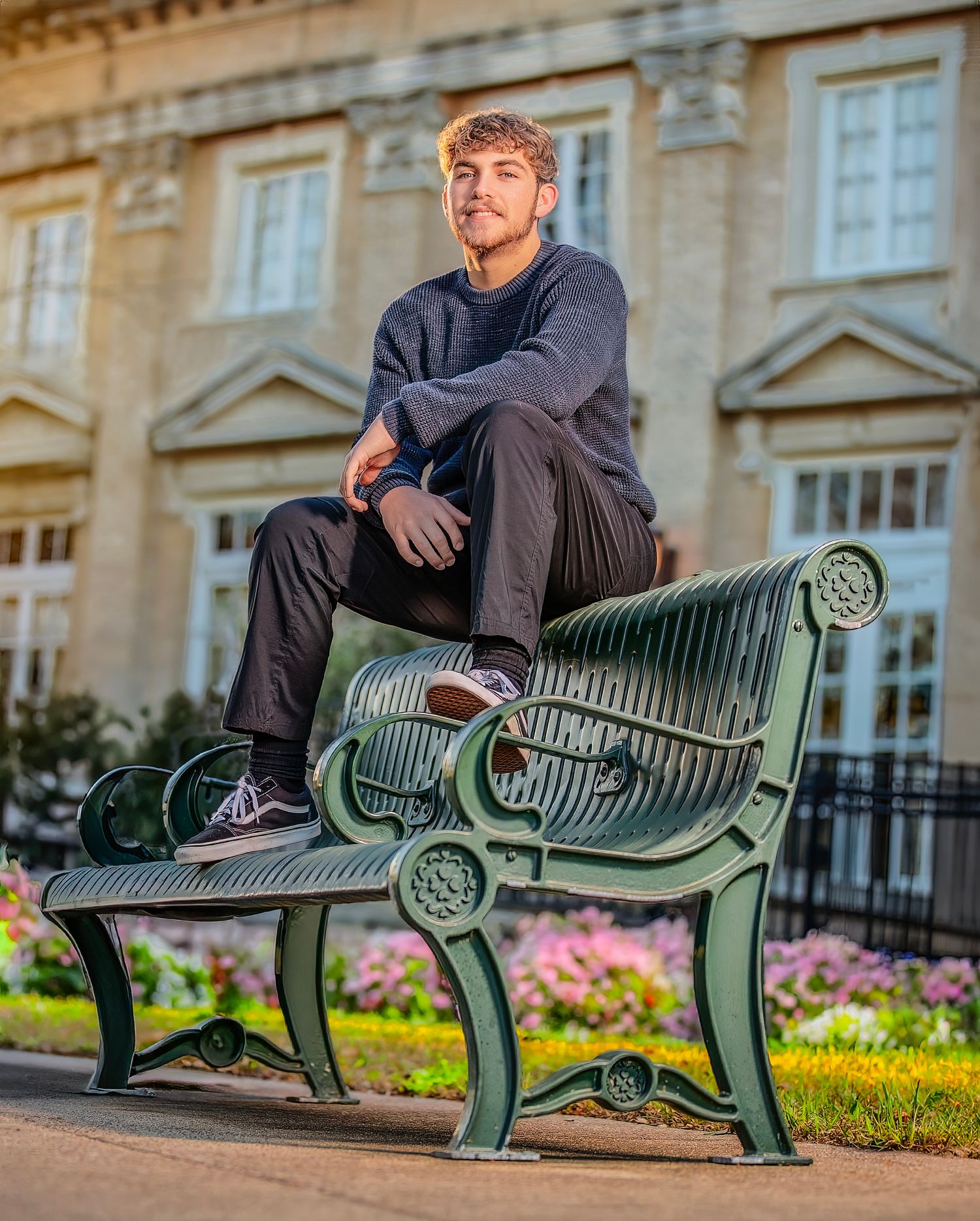 Young man sitting on a green ornate park bench in front of a grand building with colorful flower beds.