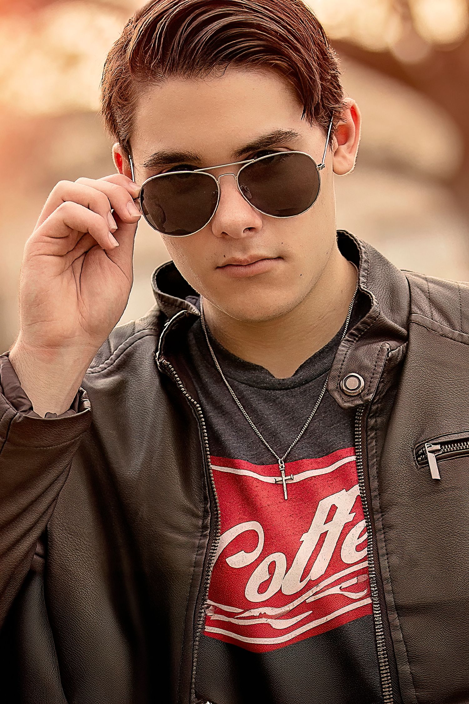 Young man in leather jacket and coffee graphic tee adjusting aviator sunglasses outdoors.
