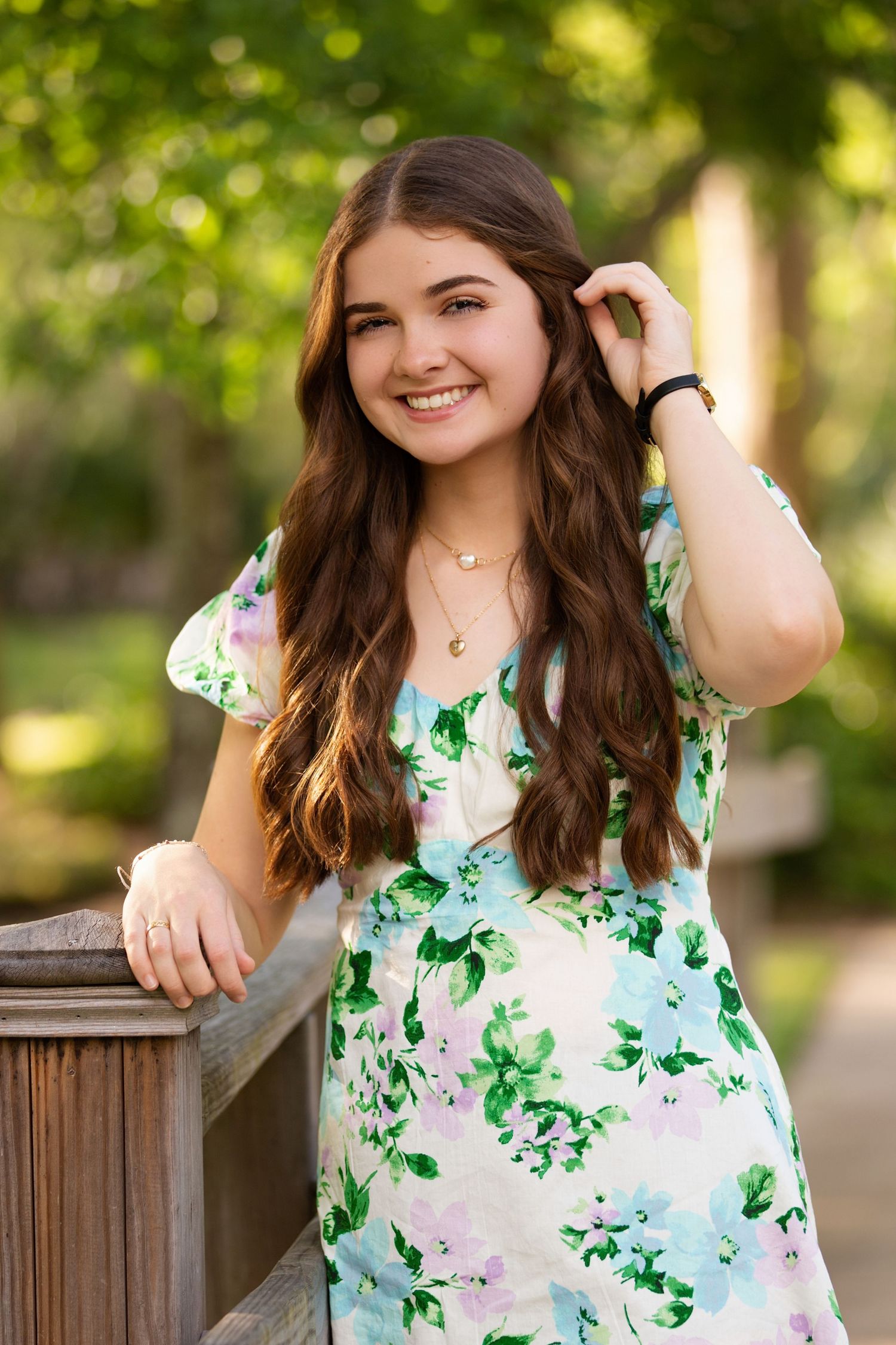 Young woman in floral dress smiling outdoors, leaning on wooden railing with lush green trees in background.