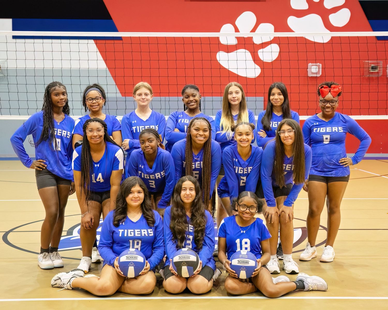 High school volleyball team in bright blue uniforms poses together on court for team photo with red wall background.