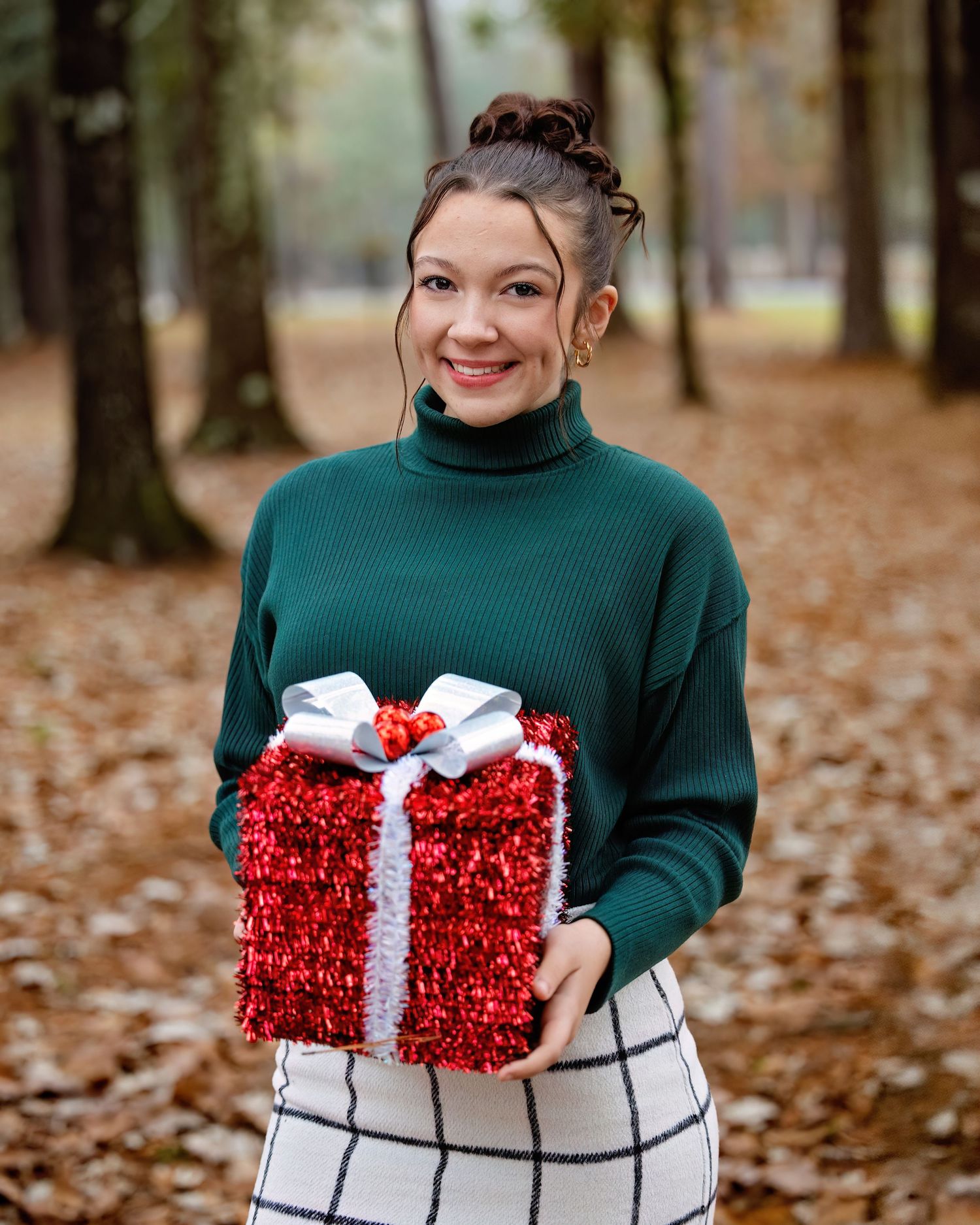 Atalyn Class of 2026,  in a dark green sweater and plaid skirt holds a sparkly red gift box with white bow against autumn leaves.