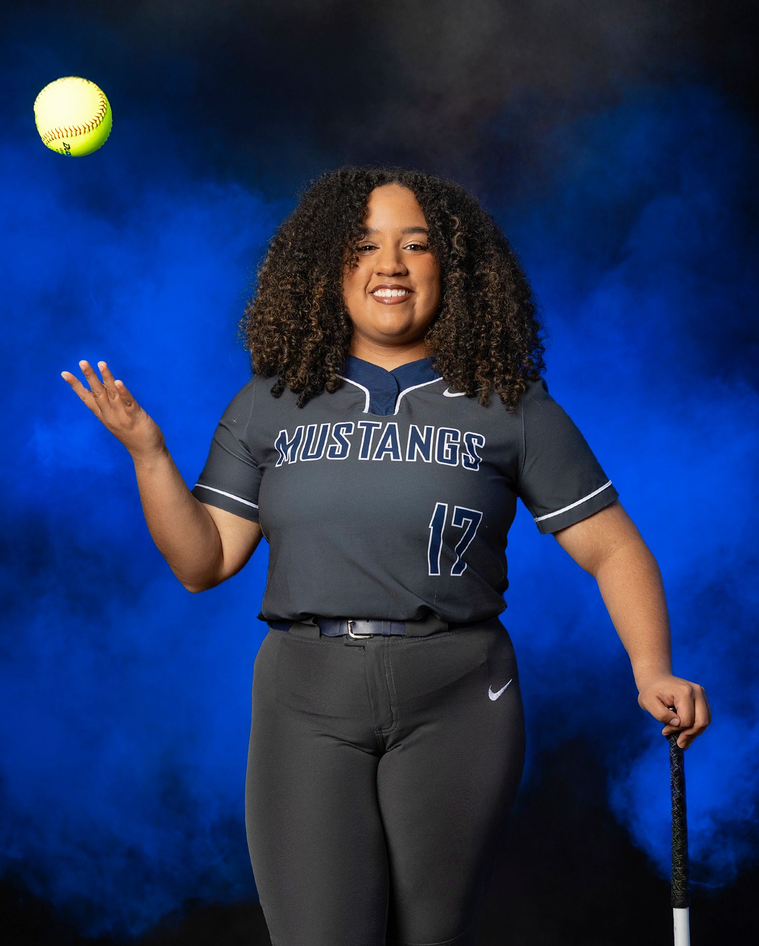 A softball player in a dark uniform throws a yellow ball against a vibrant blue background.