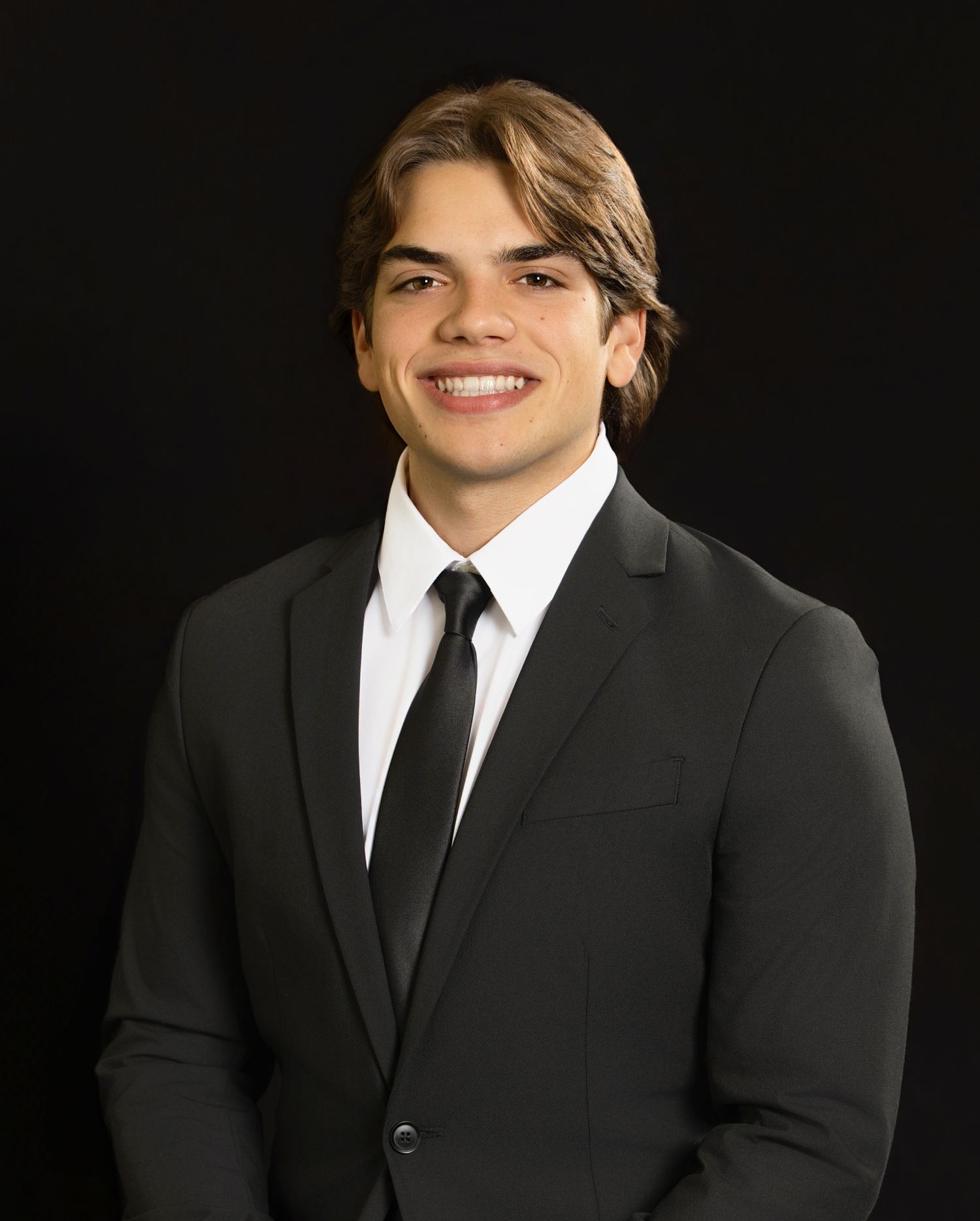 Professional headshot of someone wearing a dark suit and tie against a black background, with a bright smile.