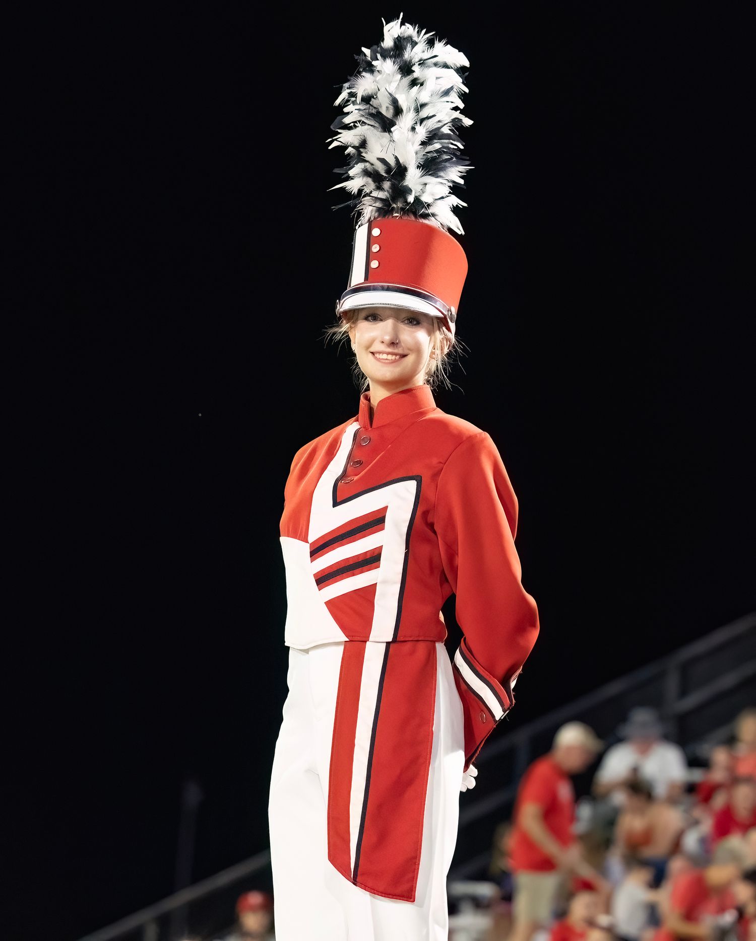 Marching band member performs in a red and white uniform with a feathered hat during a nighttime show.