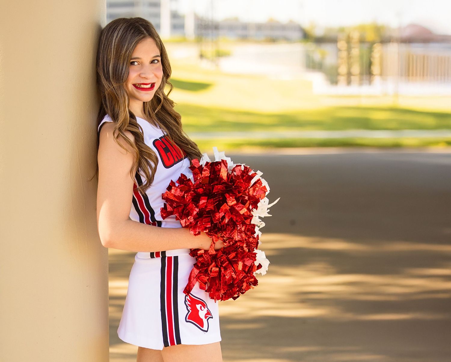 A cheerleader in a white and red uniform holds shimmering red pom poms while posing outdoors on a sunny day.