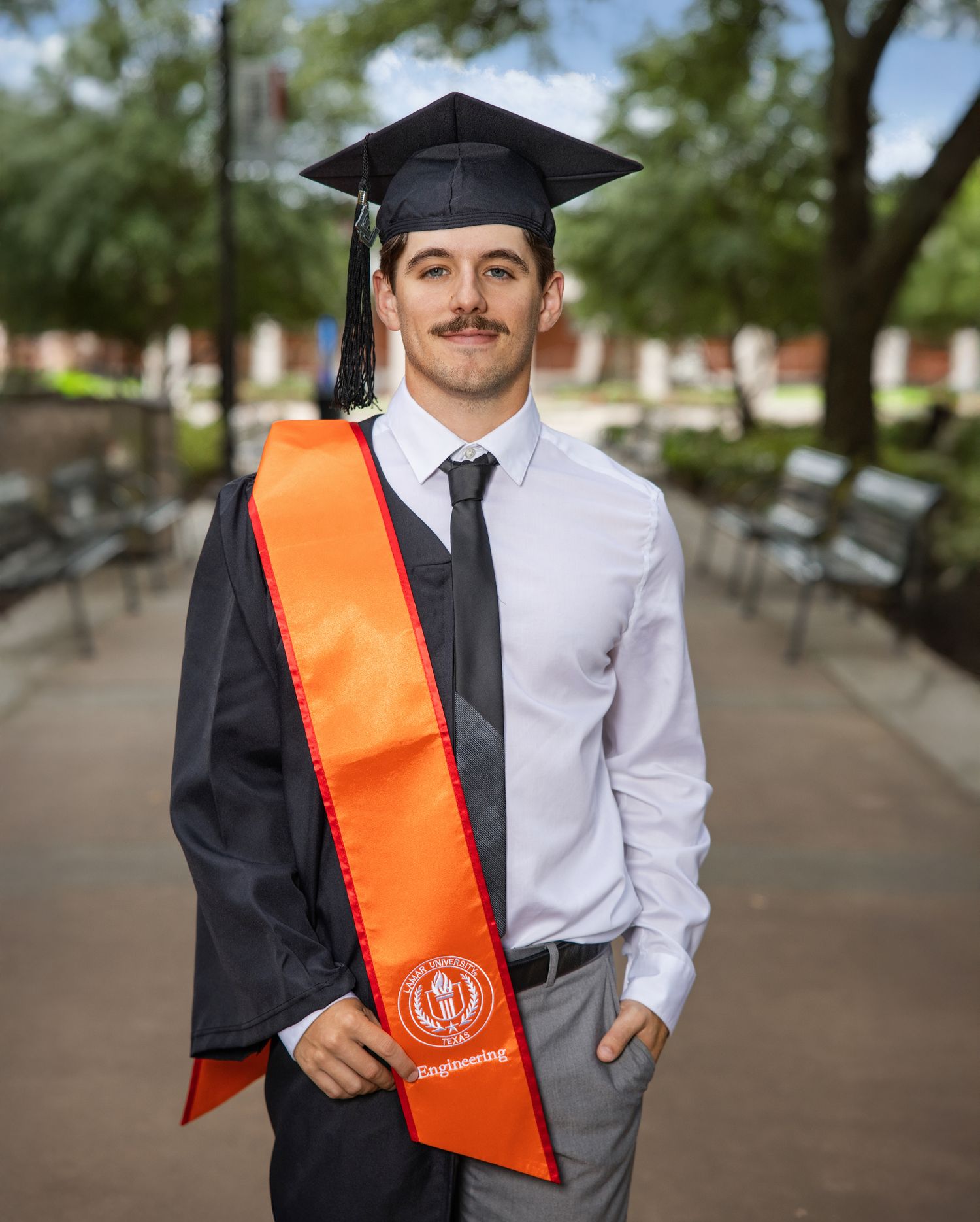 Graduate wearing black cap and gown with orange sash poses for graduation photos on campus walkway.