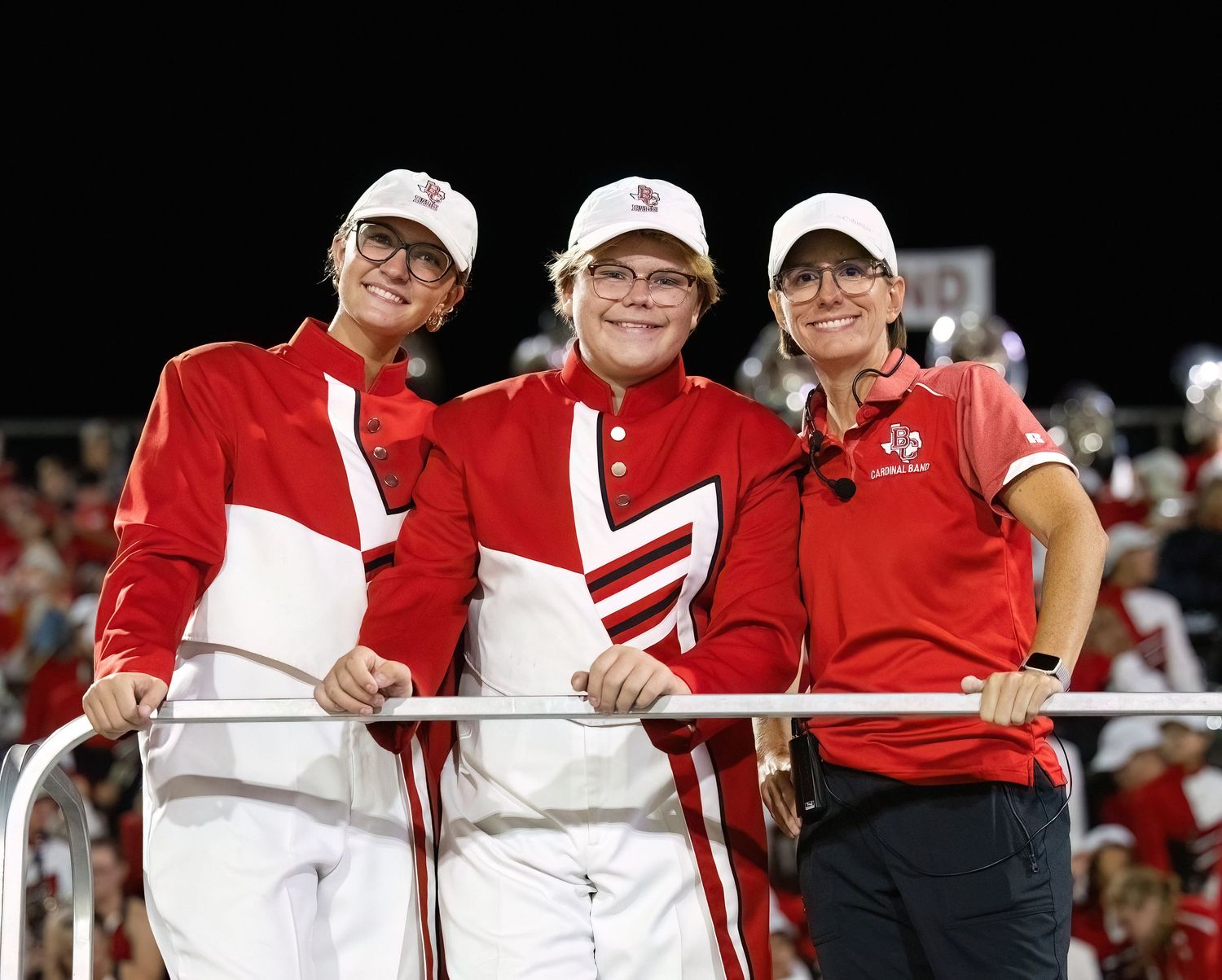 Drum Majors and director wearing red and white team colors celebrate together in the stadium stands during a game.