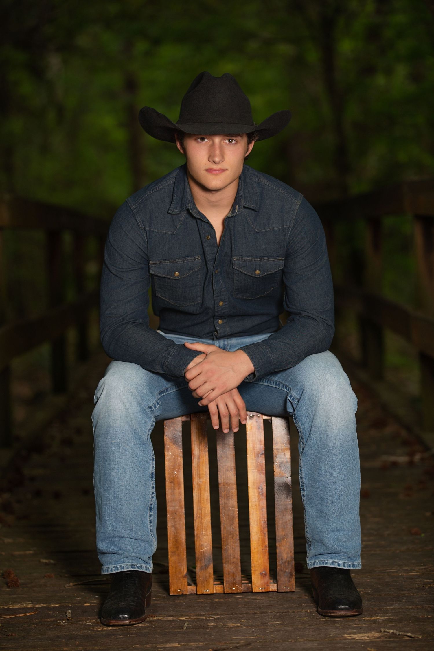Young man in black cowboy hat, denim shirt, and jeans sits on wooden crate outdoors.