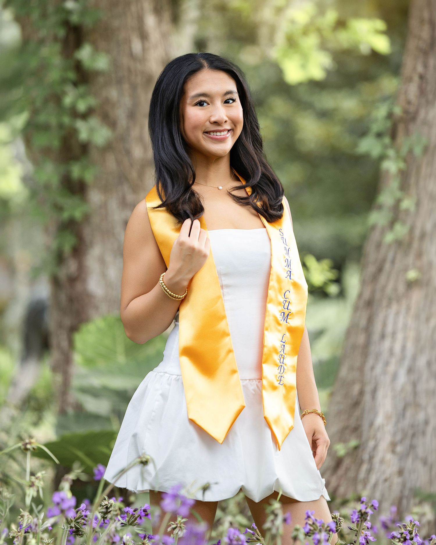 Smiling graduate in white dress with gold stole and sash poses outdoors among purple flowers and trees.