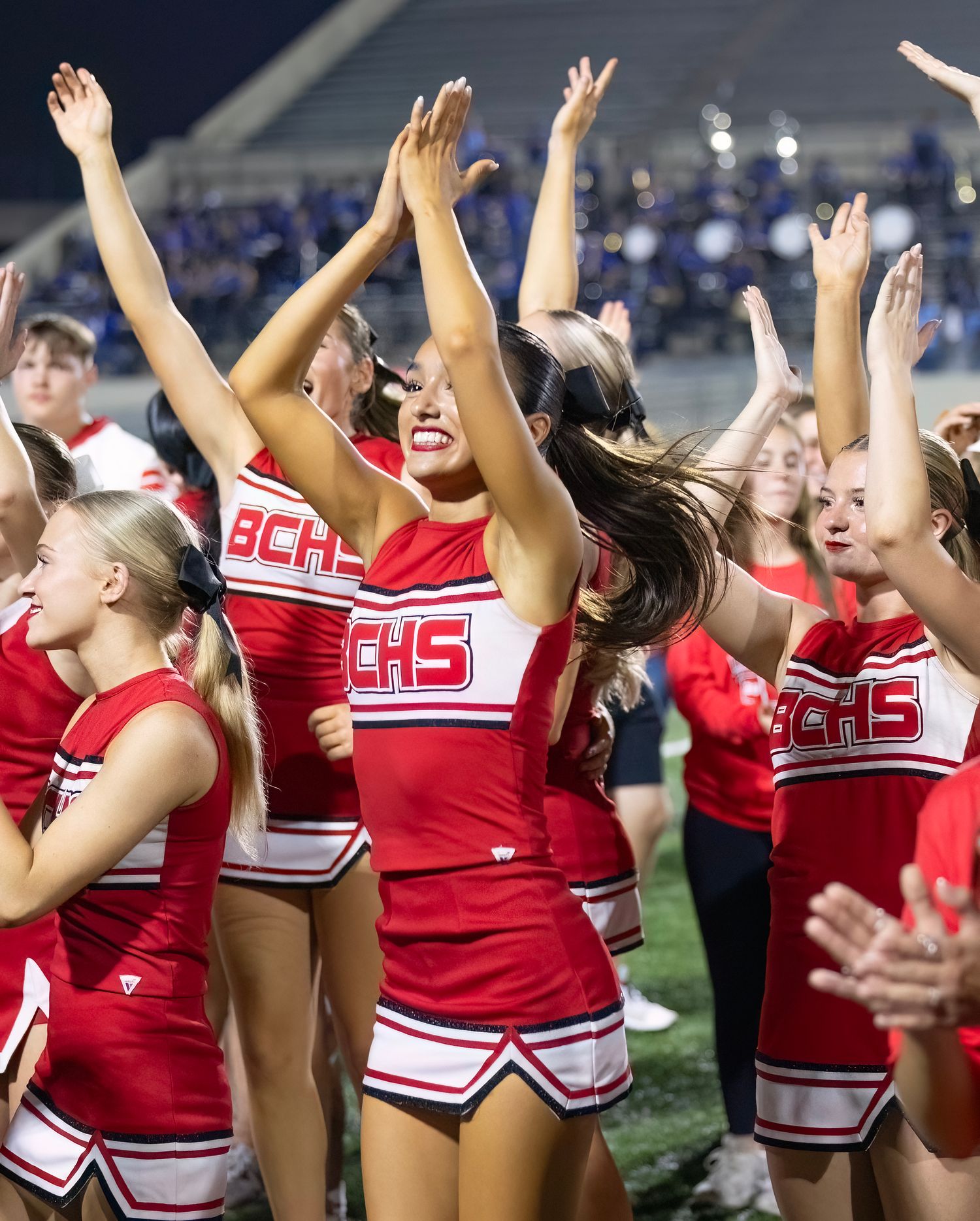 High school cheerleaders in red uniforms perform an enthusiastic celebration routine on a football field at night.