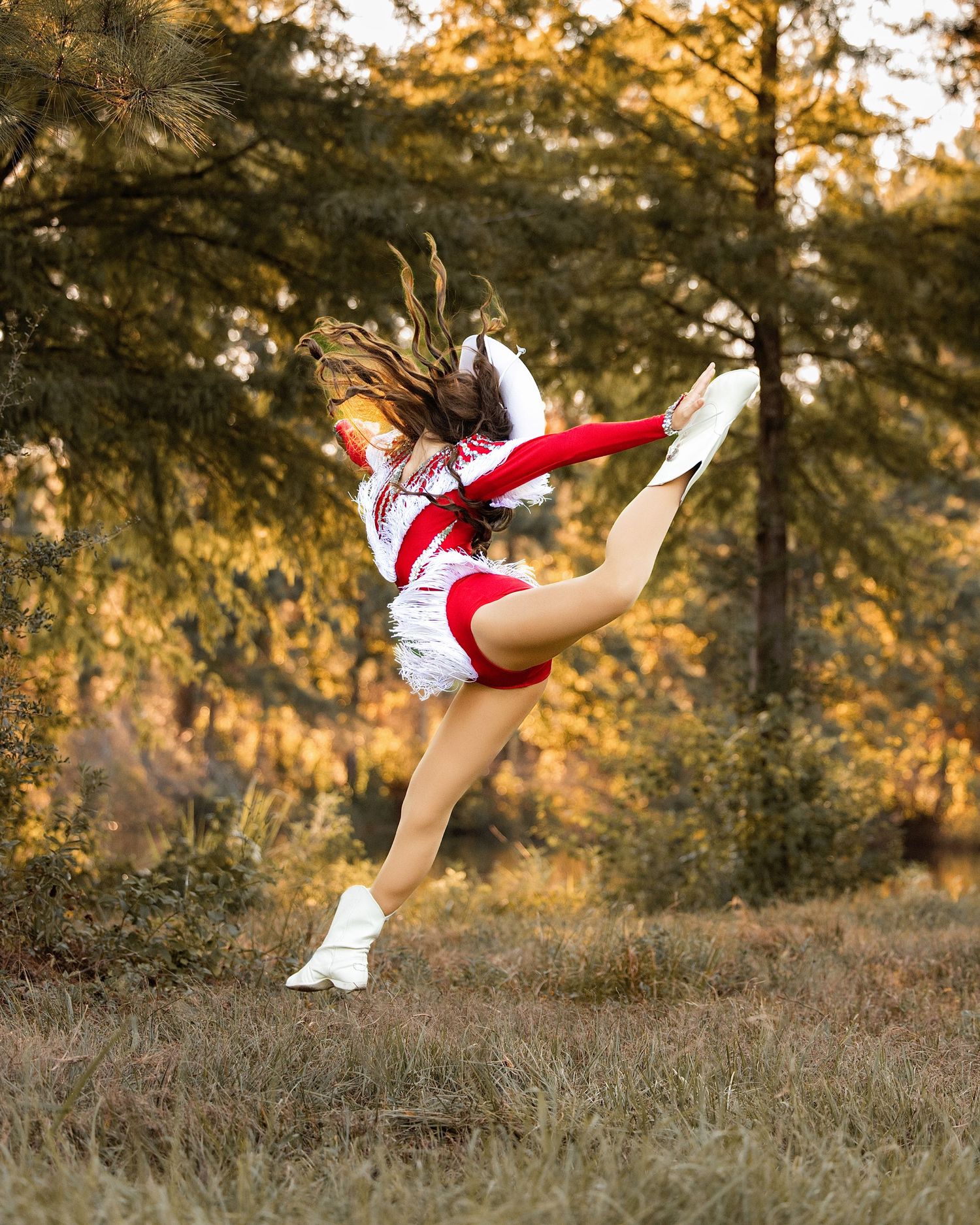A cheerleader in red and white uniform performs a high kick jump outdoors with autumn trees in the background.