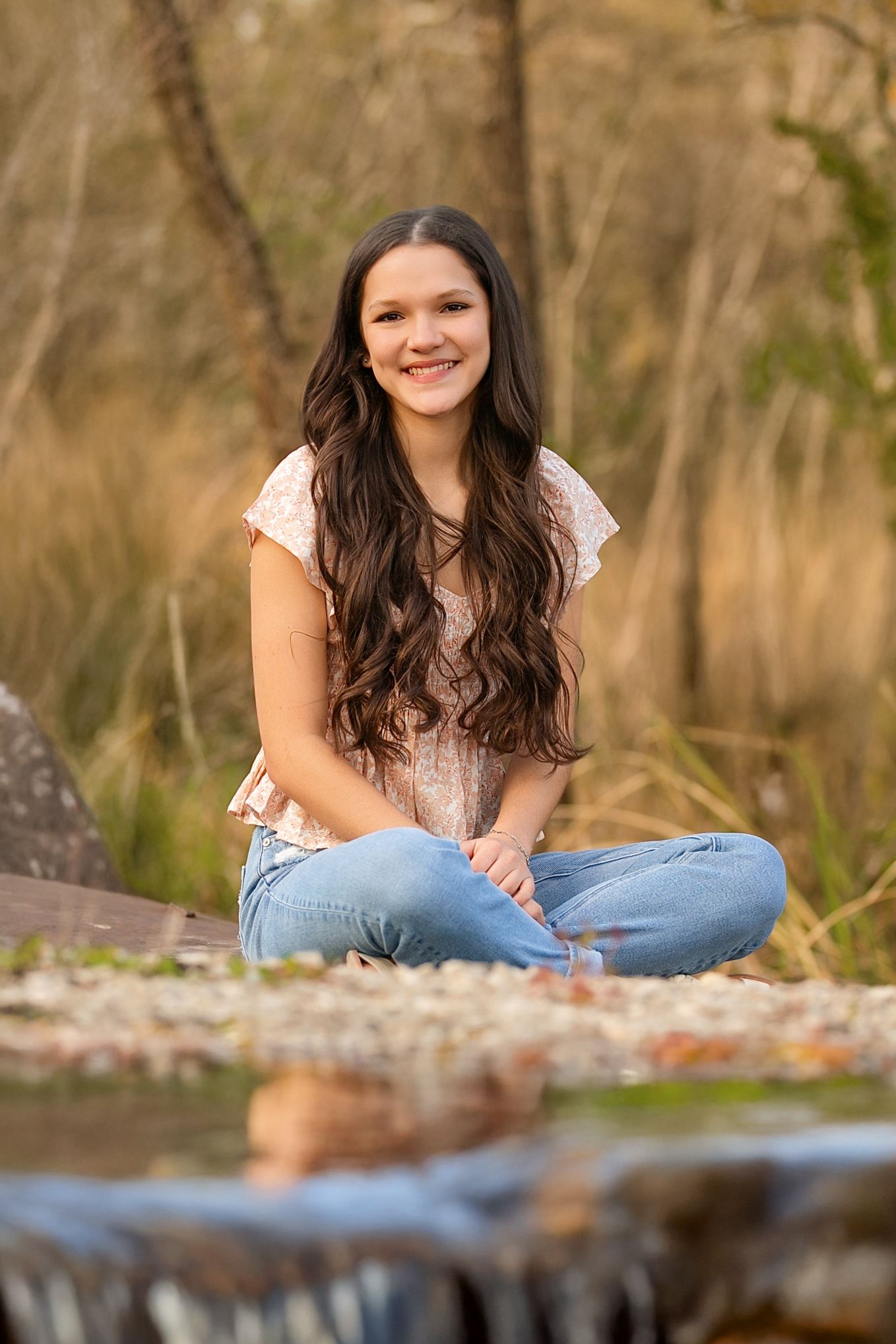 SETX Senior in a white top and jeans sits casually outdoors in a natural setting during a portrait photography session.