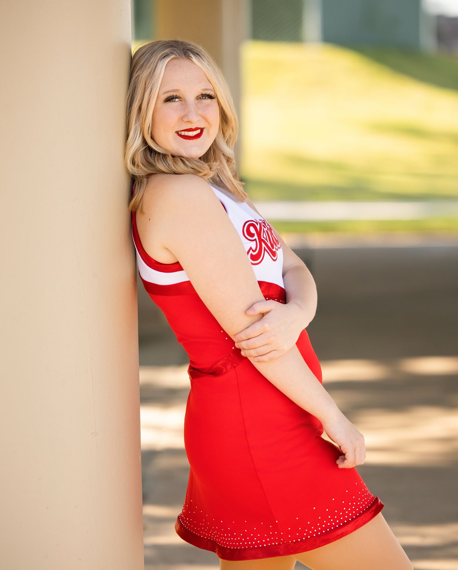 Cheerleader in red uniform poses confidently against outdoor column backdrop.