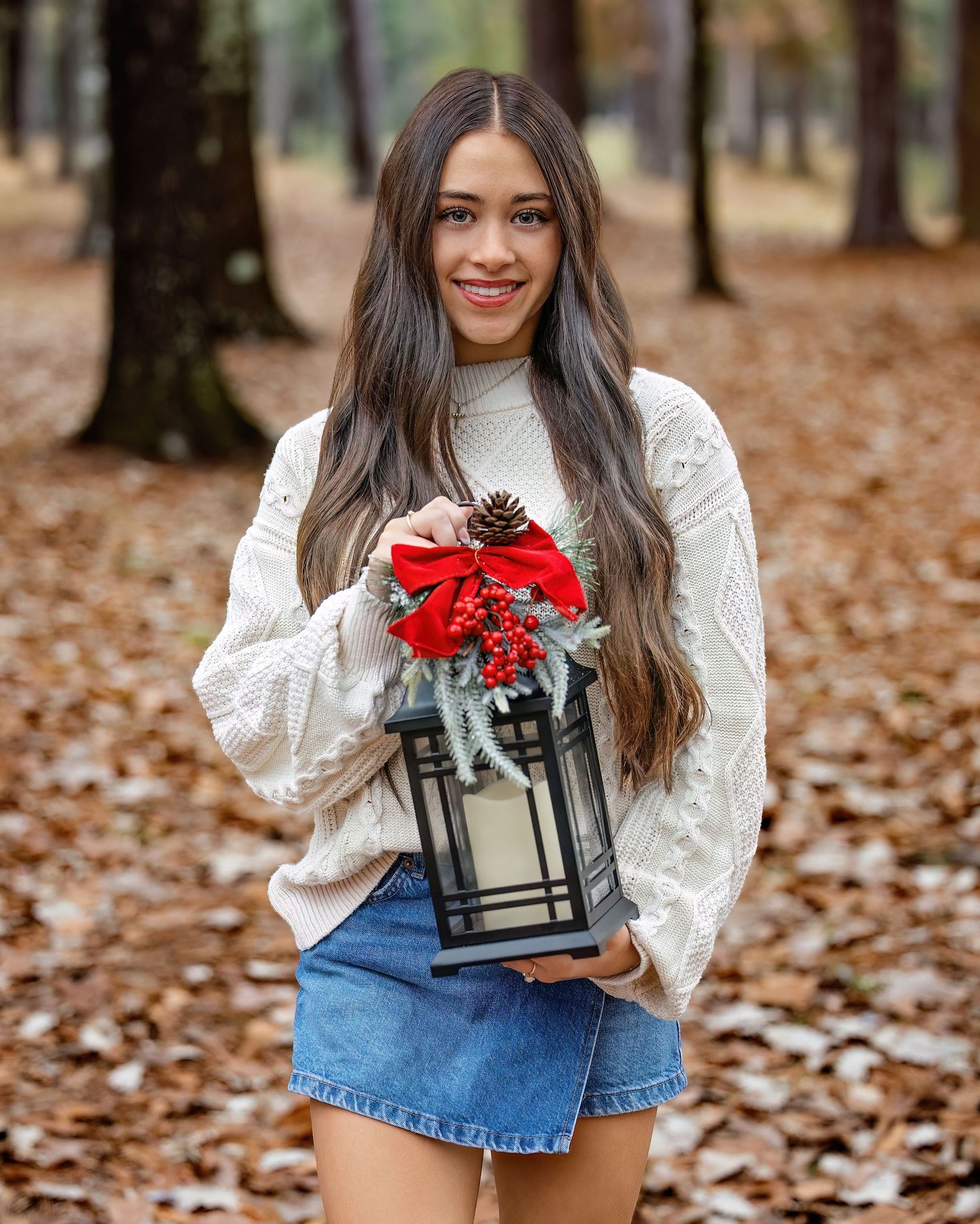 A person in a white blouse and denim skirt holds a decorative lantern with red bow among fallen autumn leaves.
