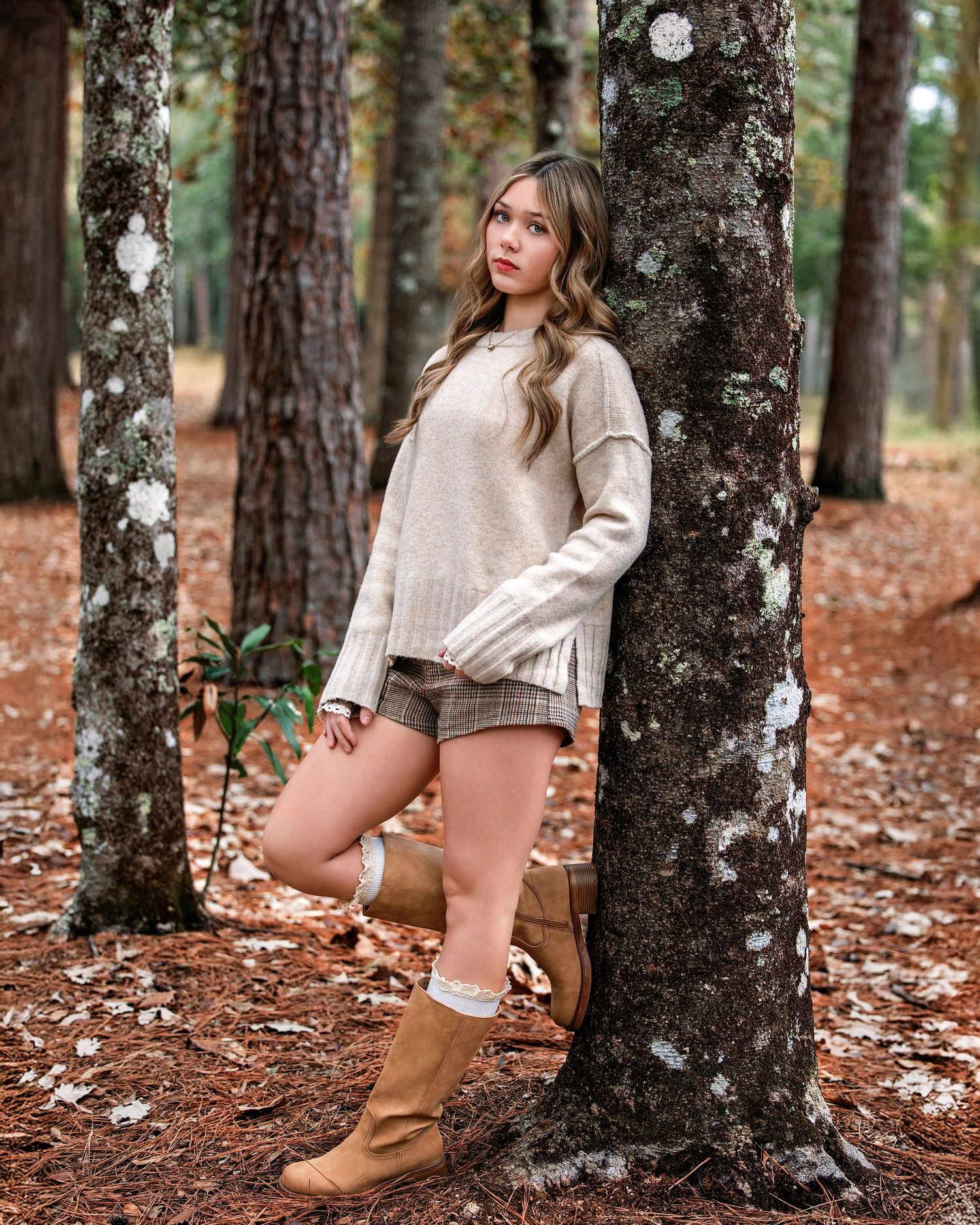 Person in beige sweater and shorts poses by tree trunks in autumn forest with fallen leaves on ground.