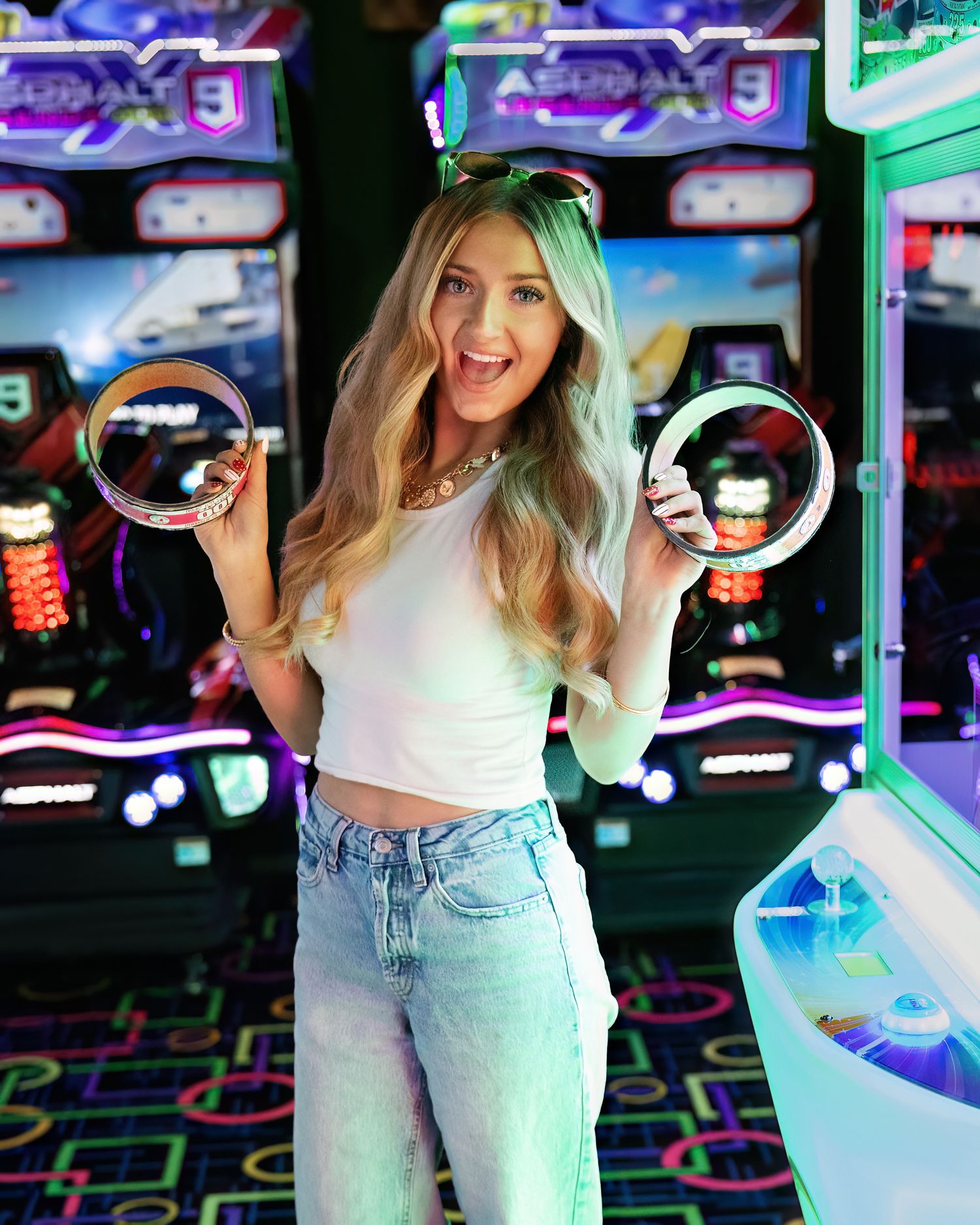 Smiling blonde woman in white crop top and jeans holding arcade game rings at a colorful neon arcade.