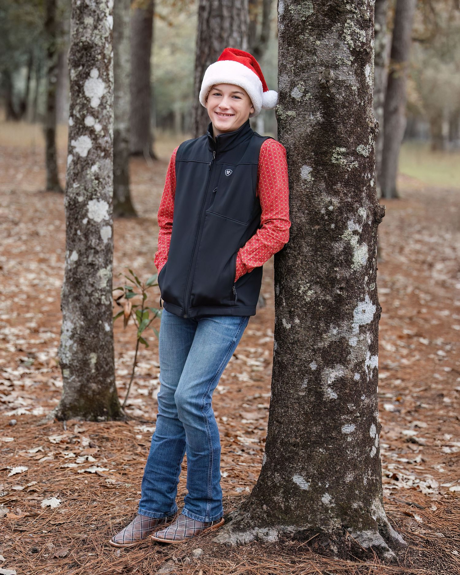 Person wearing Santa hat and winter vest poses between trees in a leaf-covered forest setting.
