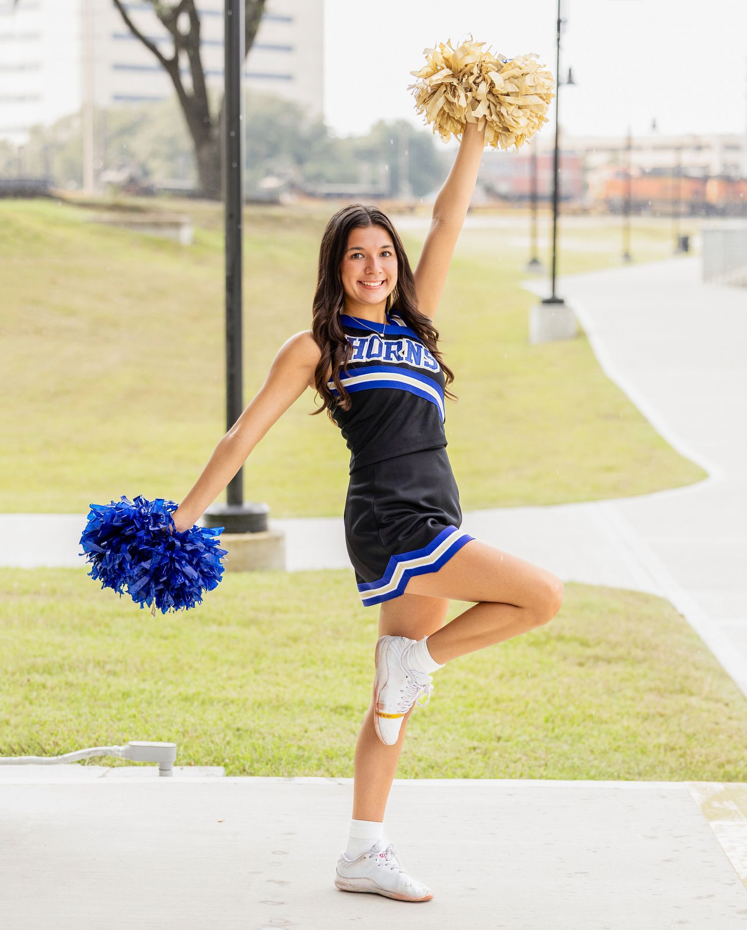 Cheerleader in blue and white uniform performs energetic pose with pom poms on campus walkway.