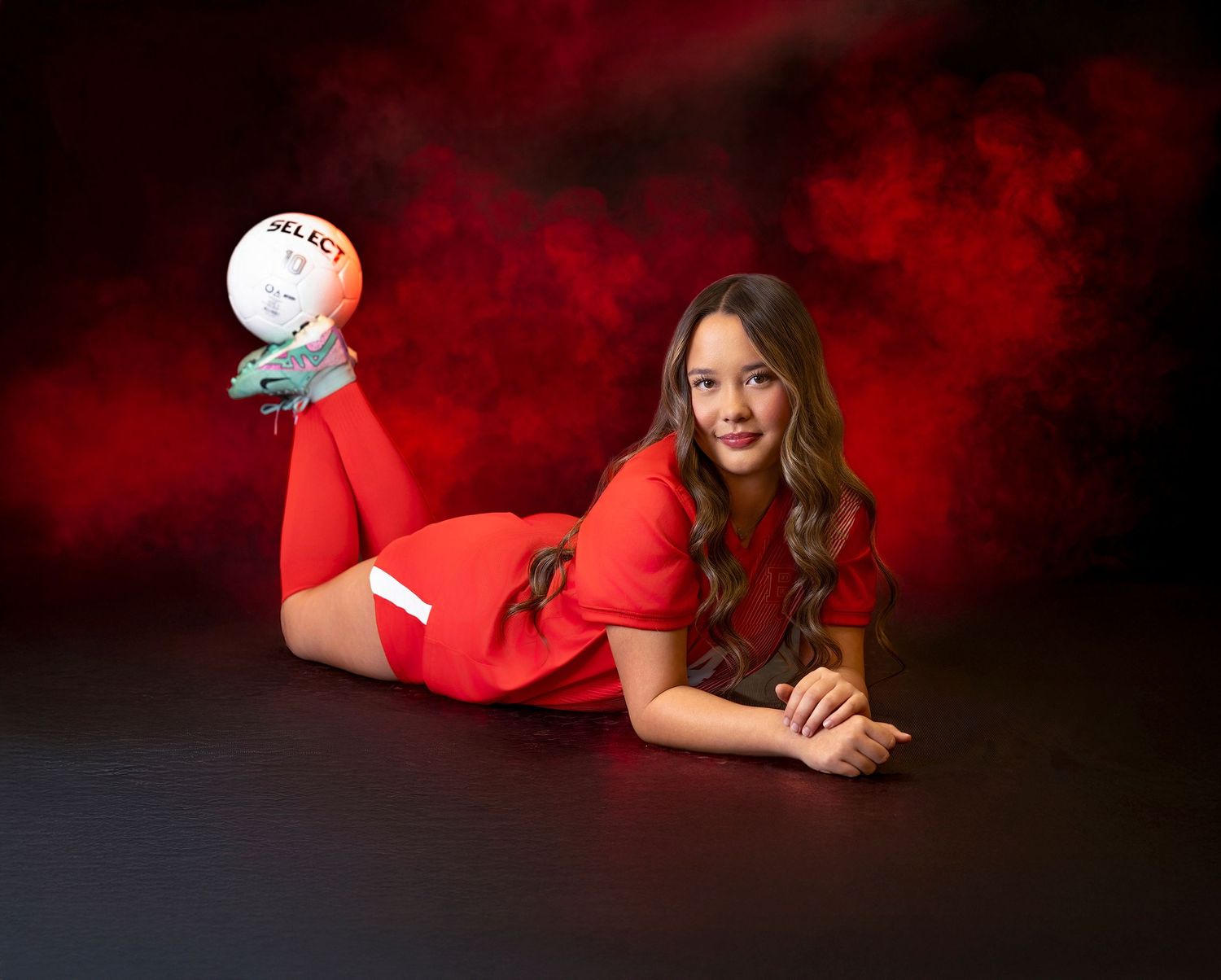 Soccer player in red uniform poses with ball against dramatic red and black background during sports photo session.