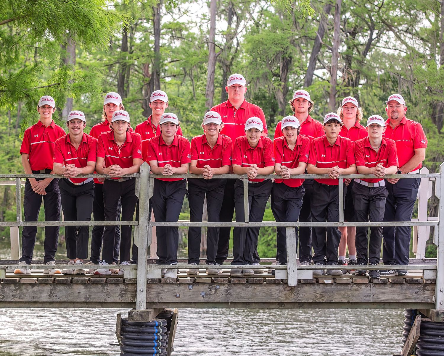 A panoramic group photo of people wearing red shirts sitting on a wooden deck in a lush green outdoor setting.