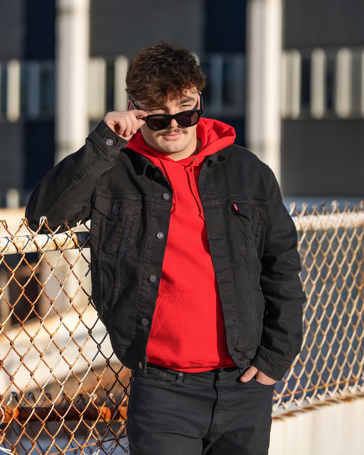 Young man in red hoodie and black denim jacket posing by chain-link fence wearing sunglasses outdoors.