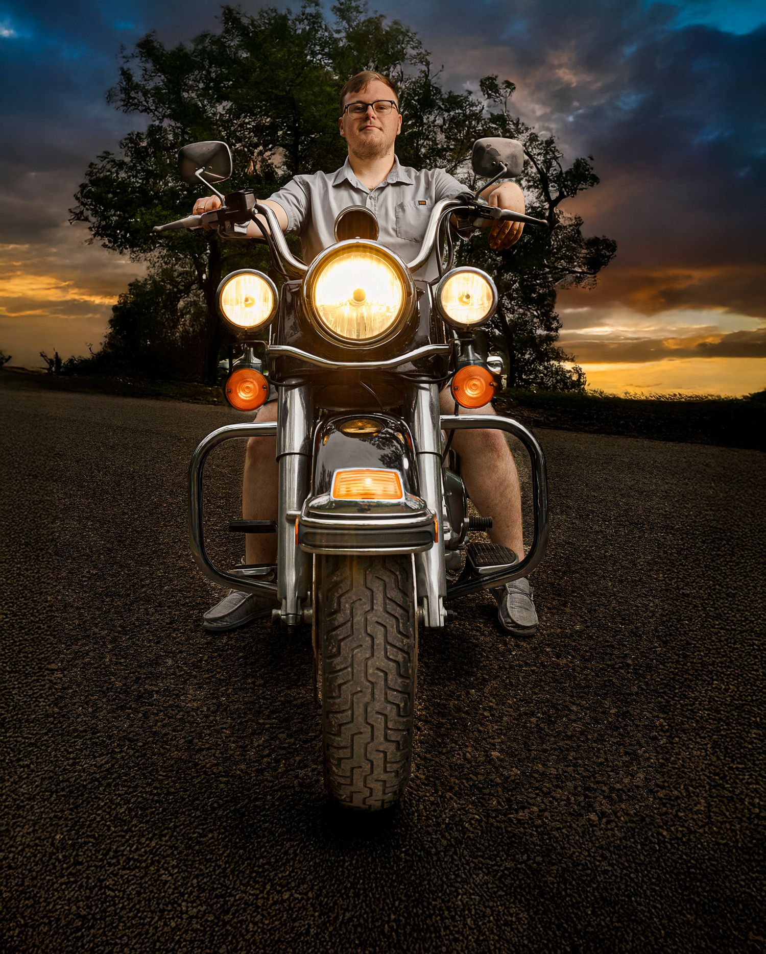 A motorcycle with illuminated headlights stands against a dramatic sunset sky with trees silhouetted in the background.