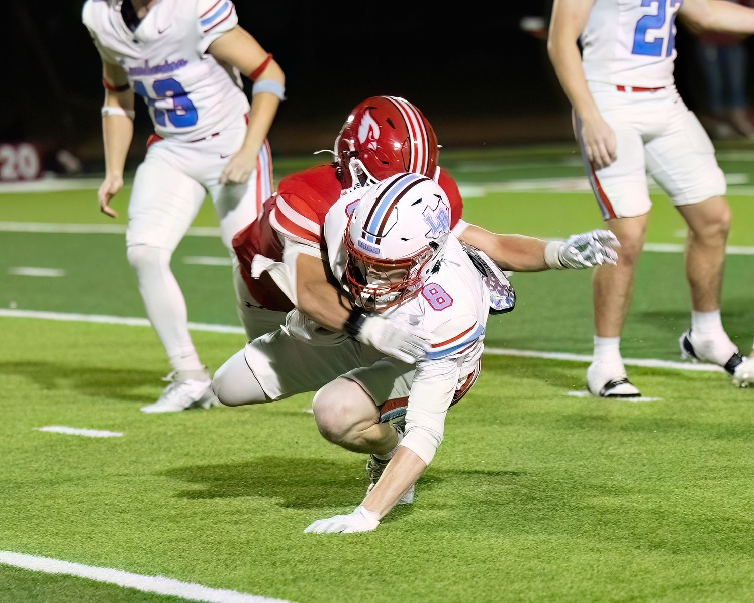 High school football player in white uniform dives forward while being tackled by player in red during night game.