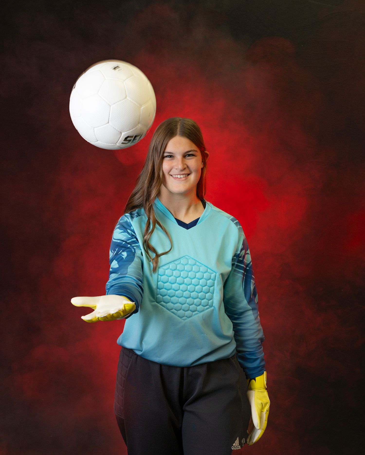 Soccer goalkeeper in light blue jersey and yellow gloves tosses white ball against dramatic red background.